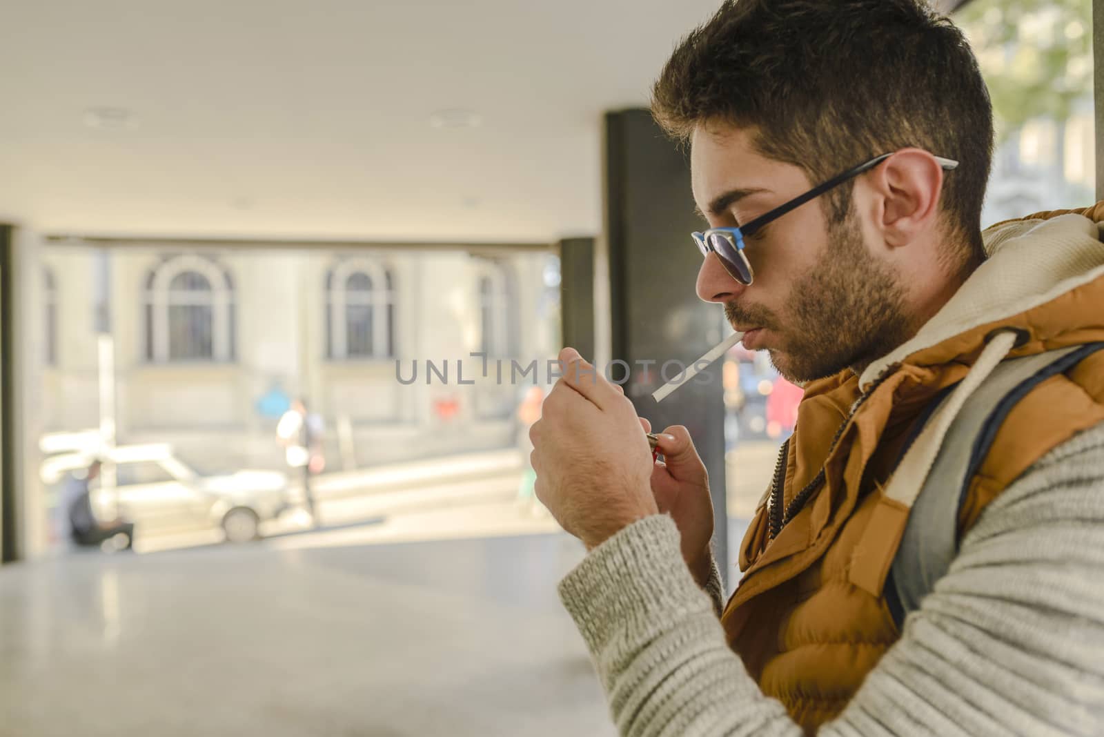 Young handsome hipster with orange jacket and sunglasses enjoying a cigarette in the street.