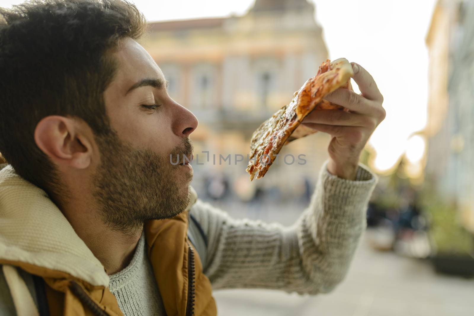 Handsome bearded young man with orange jacket eating pizza in the city street