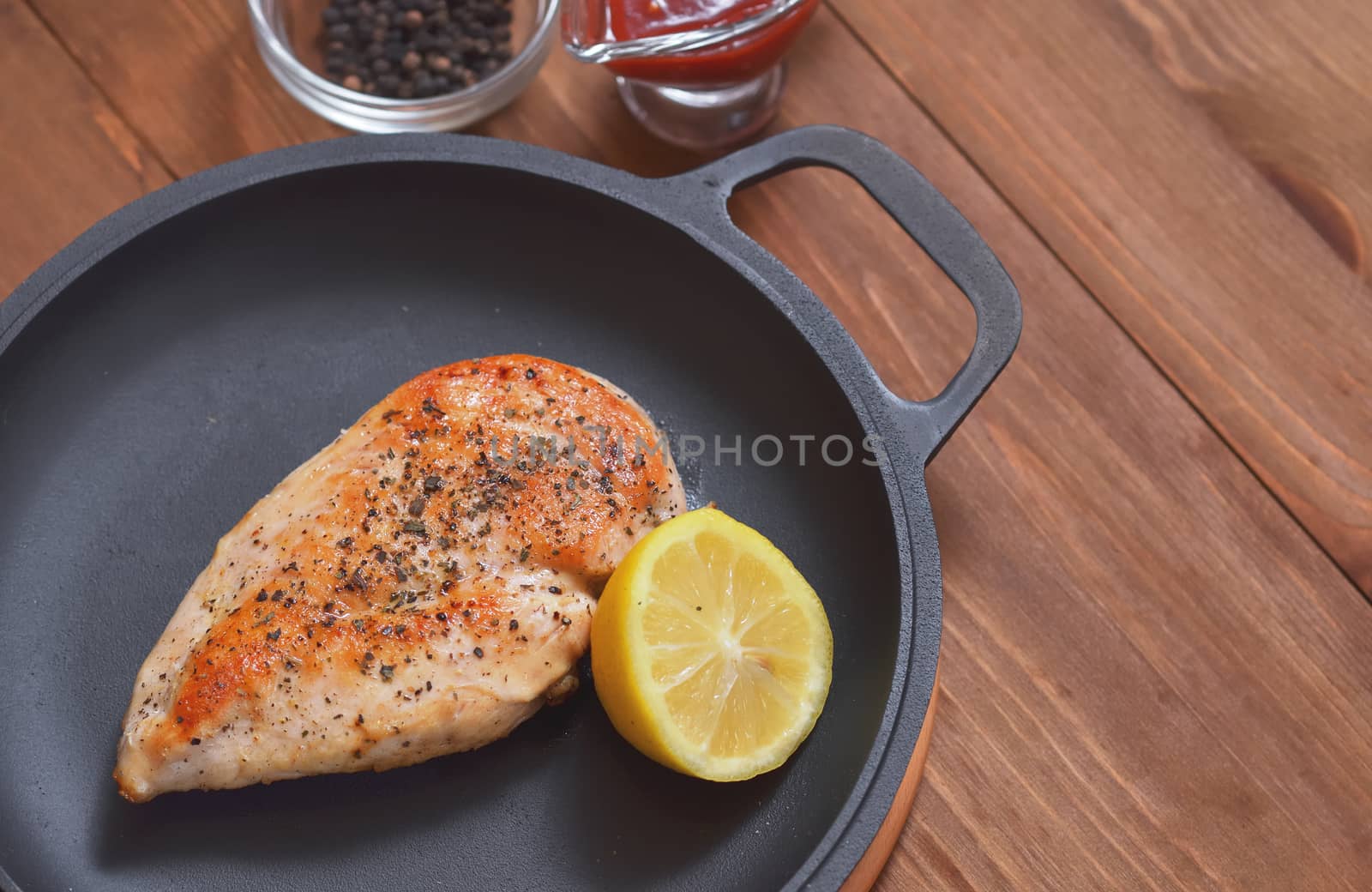 Fried chicken fillet is in a frying pan with lemon, sauce and spices on a wooden table.