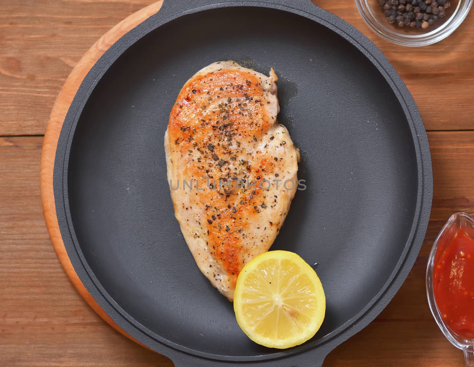 Fried chicken breast with half a lemon in a pan, spices and a sauce on a wooden table.