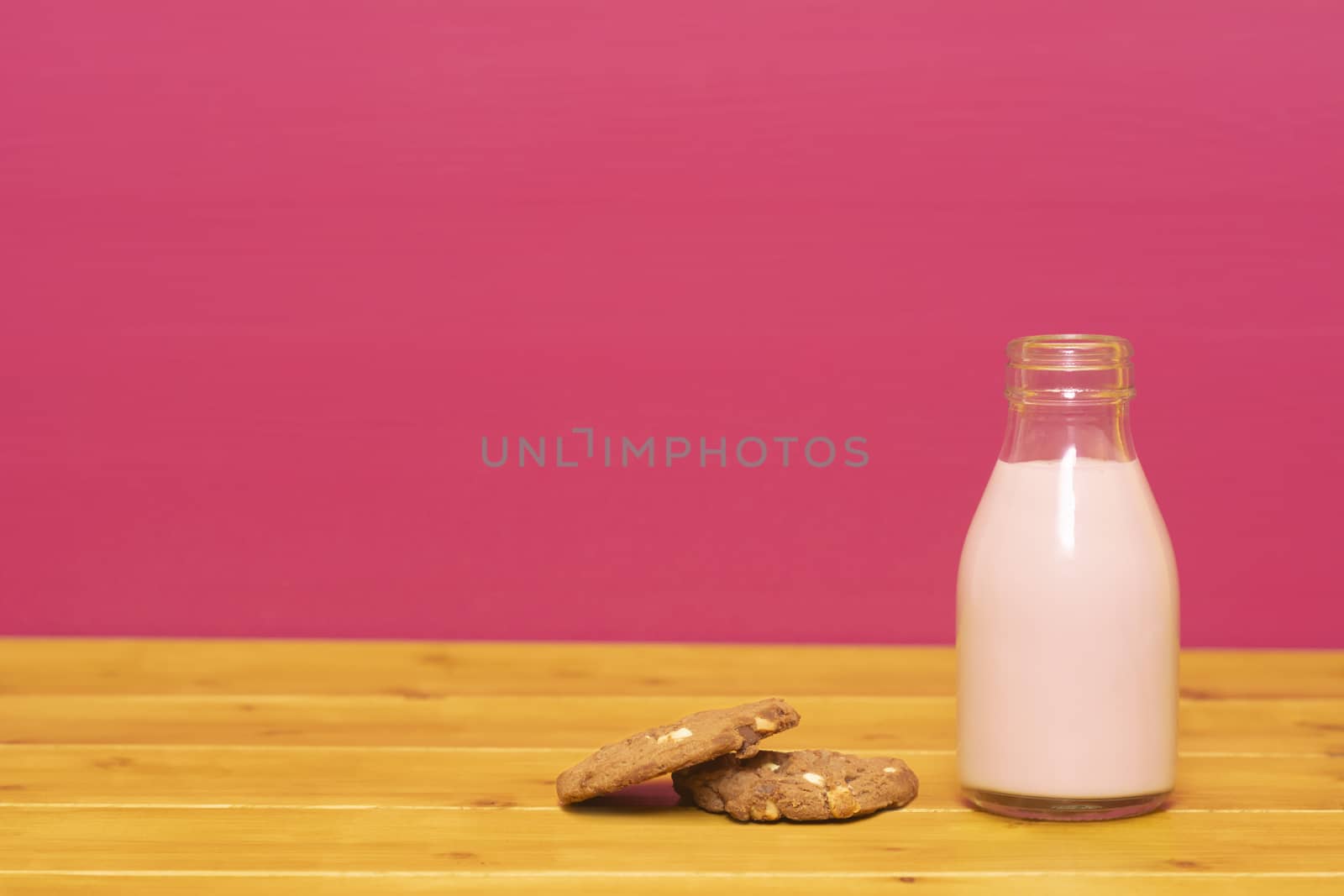 Strawberry milkshake in a one-third pint glass milk bottle and a chocolate chip cookie, on a wooden table against a pink painted background