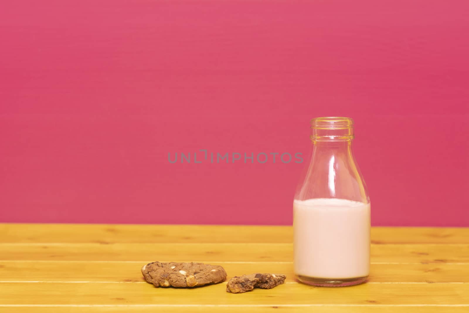 One-third pint glass milk bottle half full with strawberry milkshake and a half-eaten chocolate chip cookie, on a wooden table against a pink background