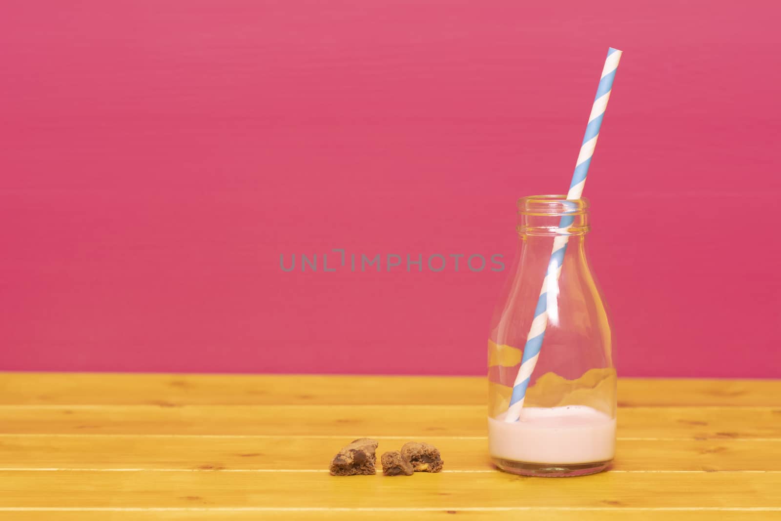 One-third pint glass milk bottle half full with strawberry milkshake with a retro straw and chocolate chip cookie crumbs, on a wooden table against a pink background