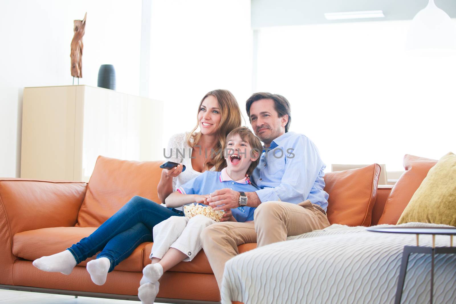 Beautiful young parents and their son are watching TV, eating popcorn and smiling while sitting on couch at home