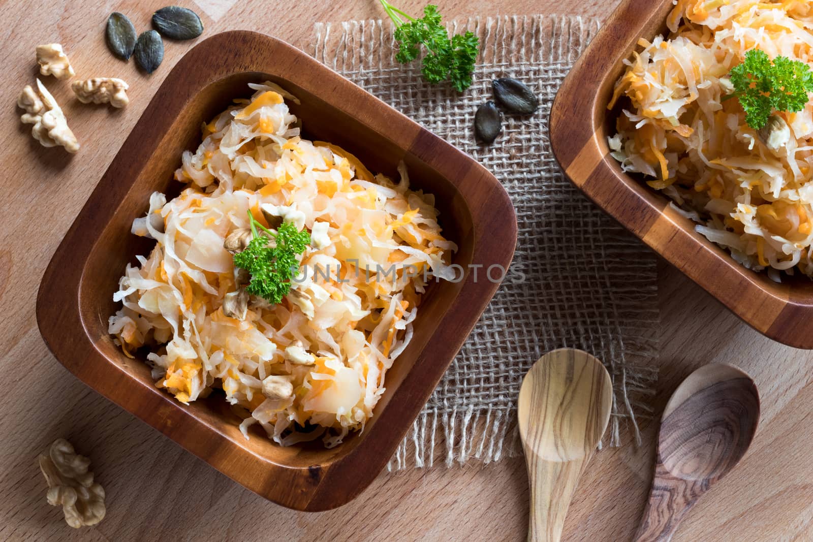 Top view of fermented cabbage and carrots in two wooden bowls on a wooden table, with walnuts, parsley, pumpkin seeds and two spoons in the background