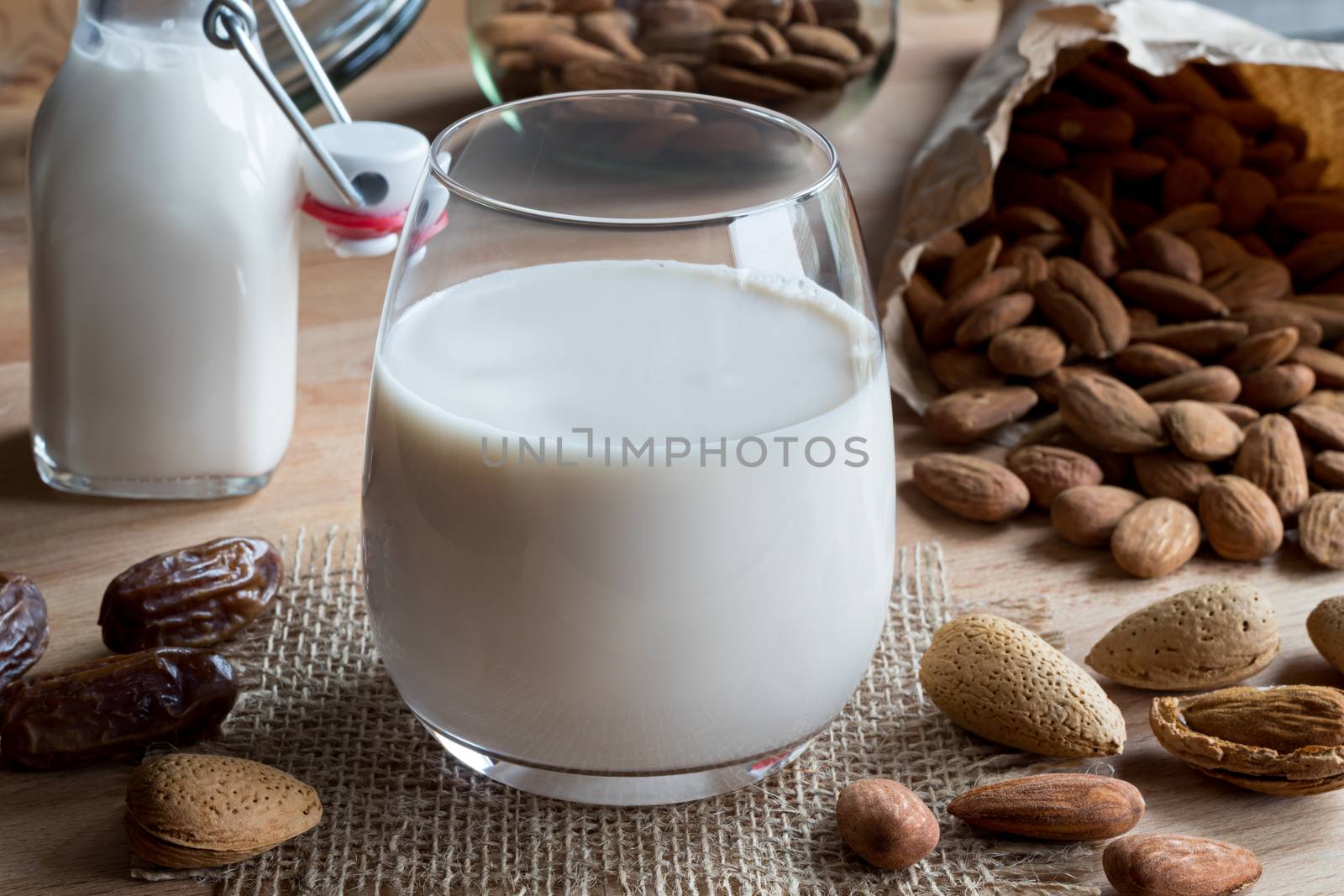 A glass of almond milk on a wooden table, with shelled, unshelled and soaked almonds and dates in the background