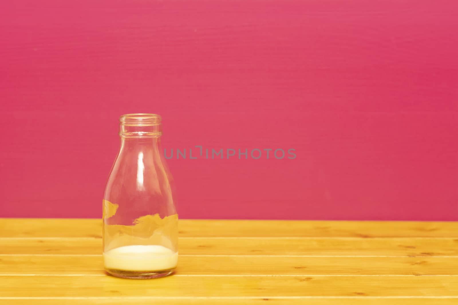 One-third pint glass milk bottle with dregs of banana milkshake, on a wooden table against a pink painted background