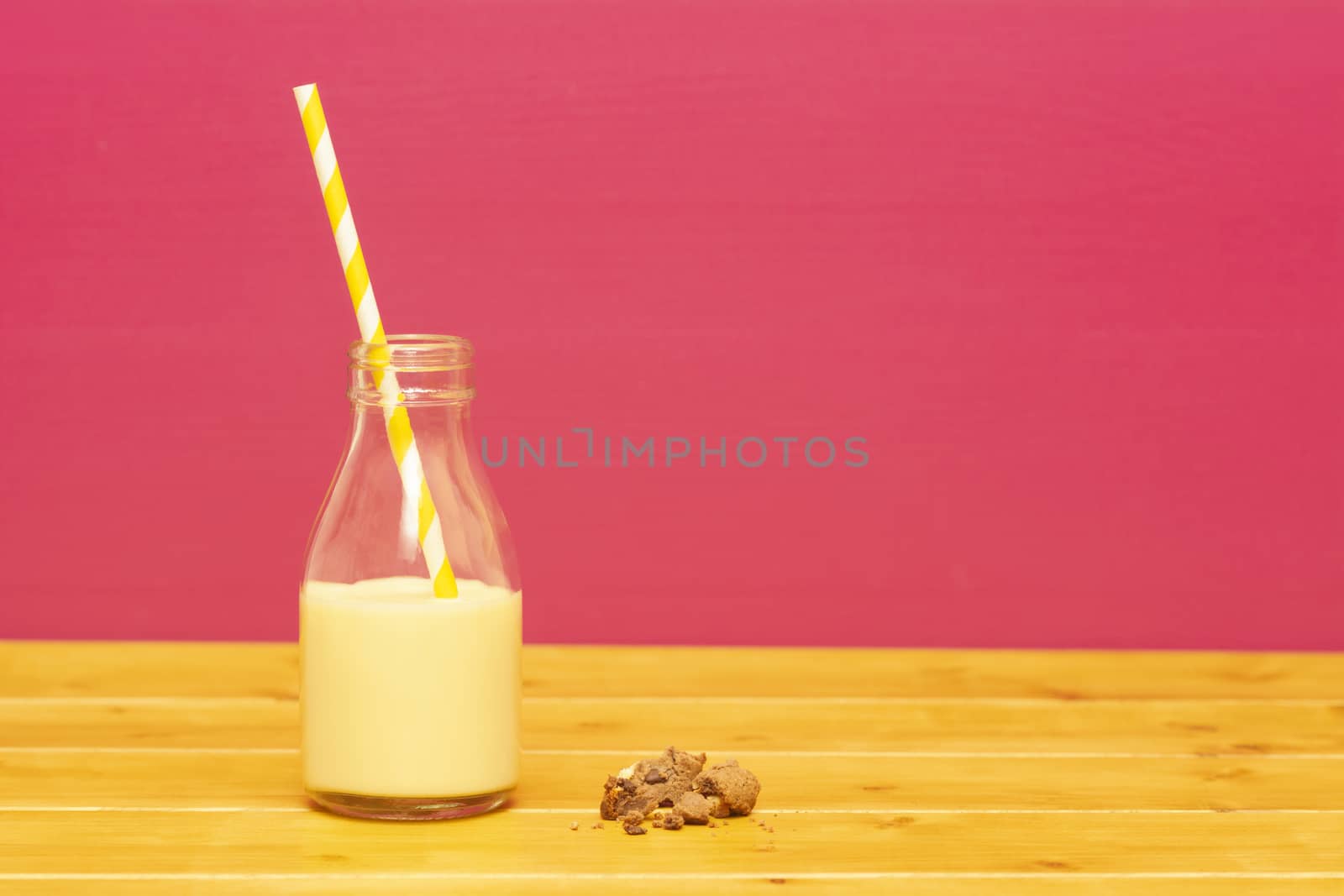 One-third pint glass milk bottle half full with banana milkshake with a retro straw and a half-eaten chocolate chip cookie, on a wooden table against a pink background