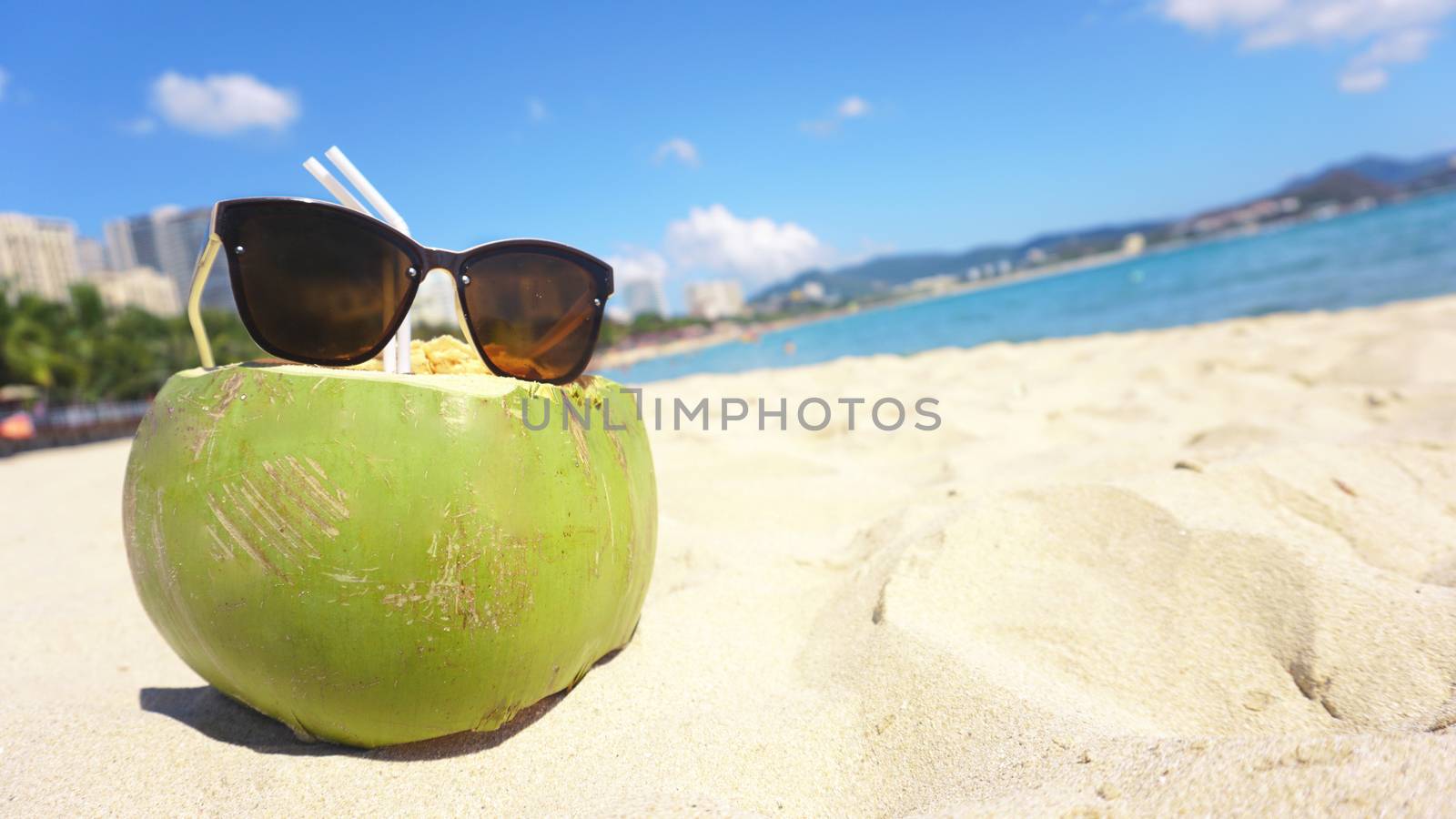 Green funny coconut in sun glasses on white sandy beach, summer travel concept background.
