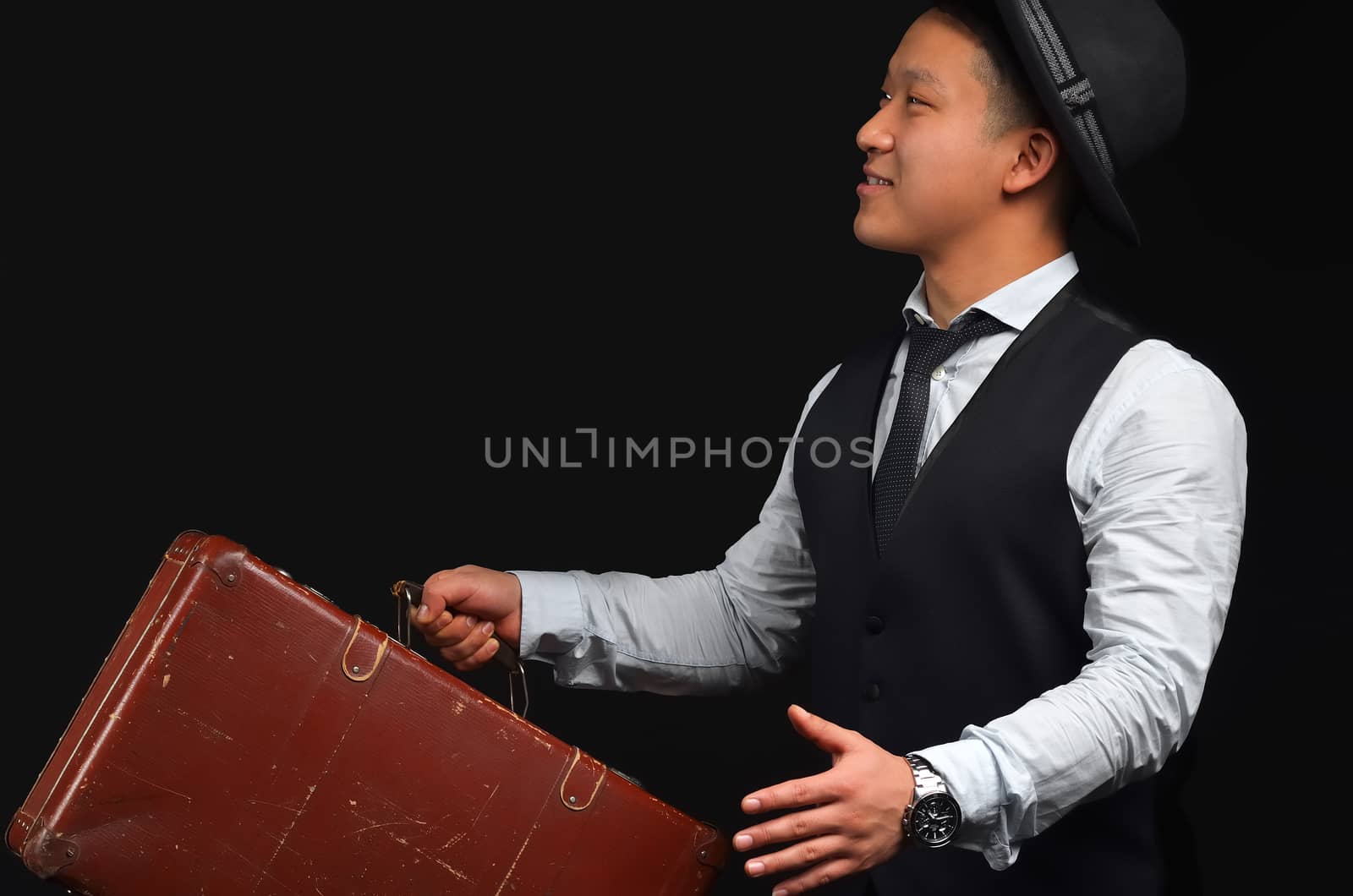 Asian young man wearing a hat with a suitcase, smiling and lifting his arms looking sideways