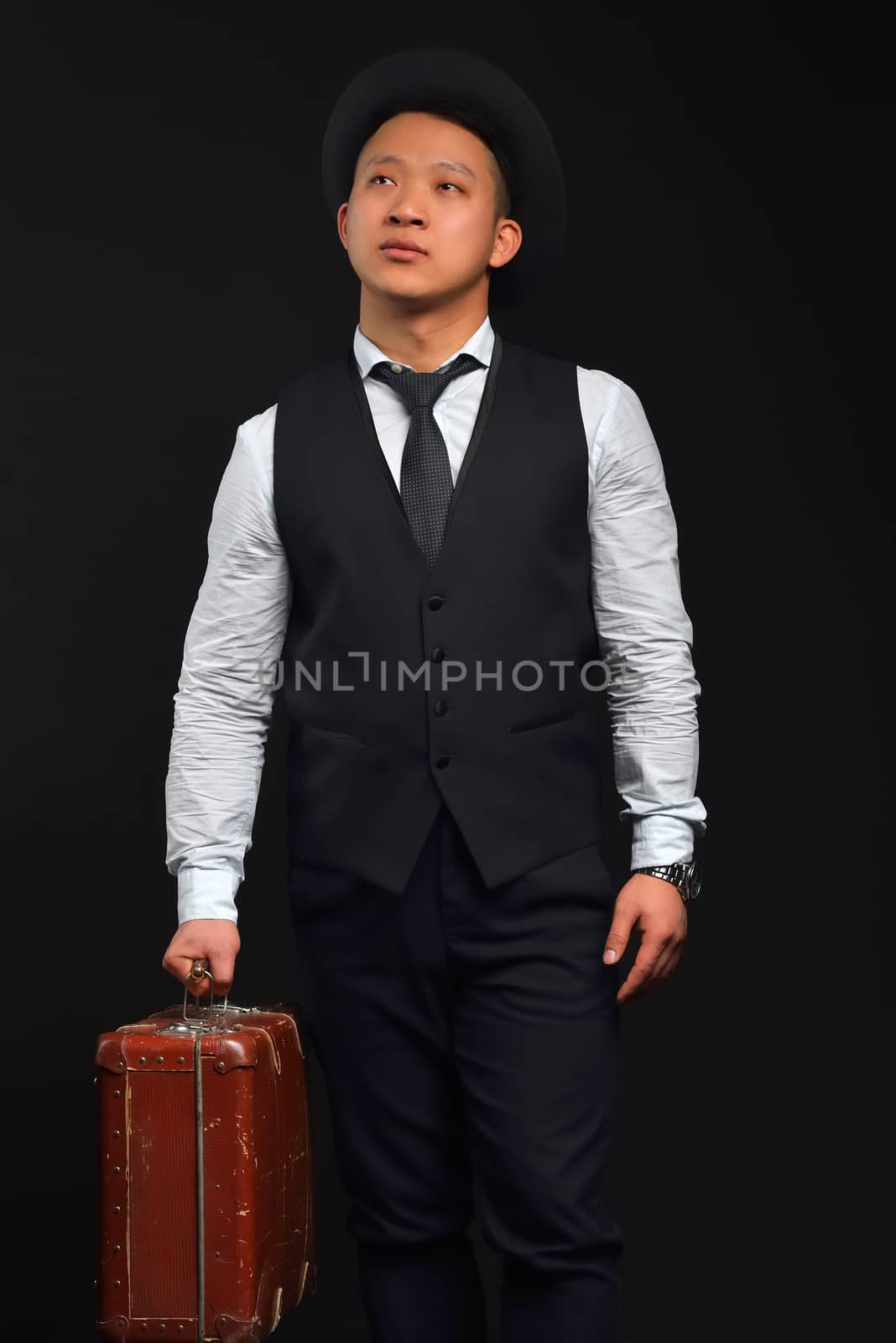 Asian man in a hat and a suit with a brown suitcase looks up, thoughtful, photo in full swing