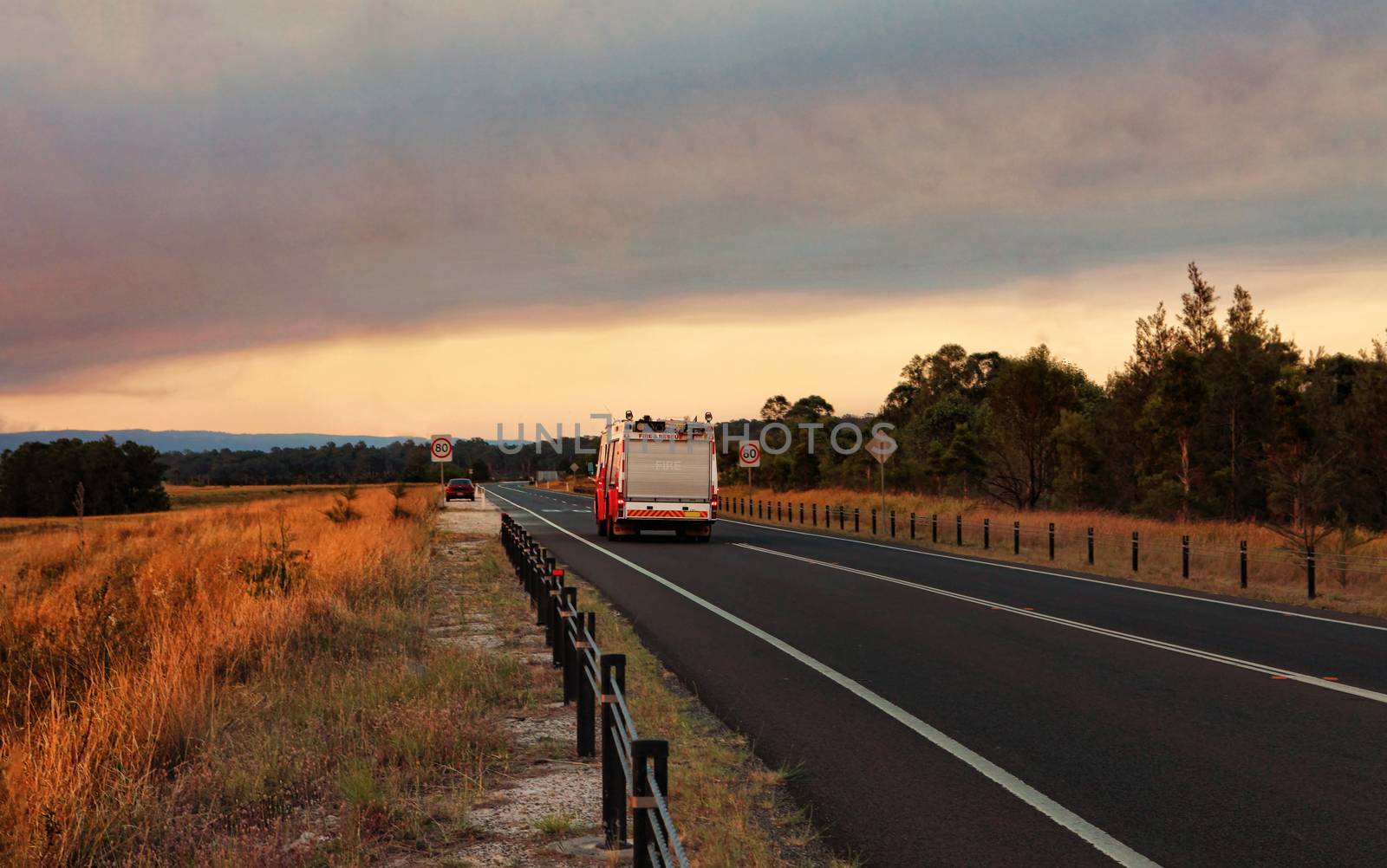 A  Rural Fire Service, fire and rescue vehicle responds urgently to a large out of control bushfire in the mountains.  Smoke and sun casts a red glow across the landscape.  Thick smoke billowing overhead