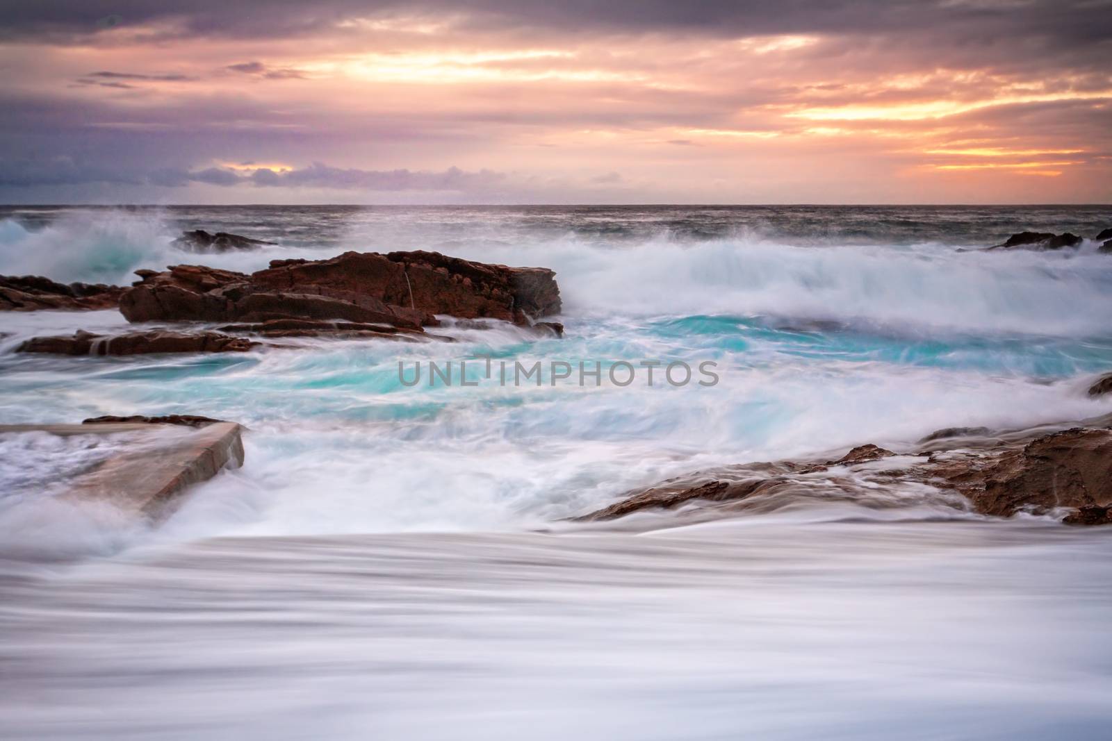 Large swell brings in big waves that create fast flowing overflows at the ocean rock pool. The glow of the morning sun as it trys to break free of through the clouds