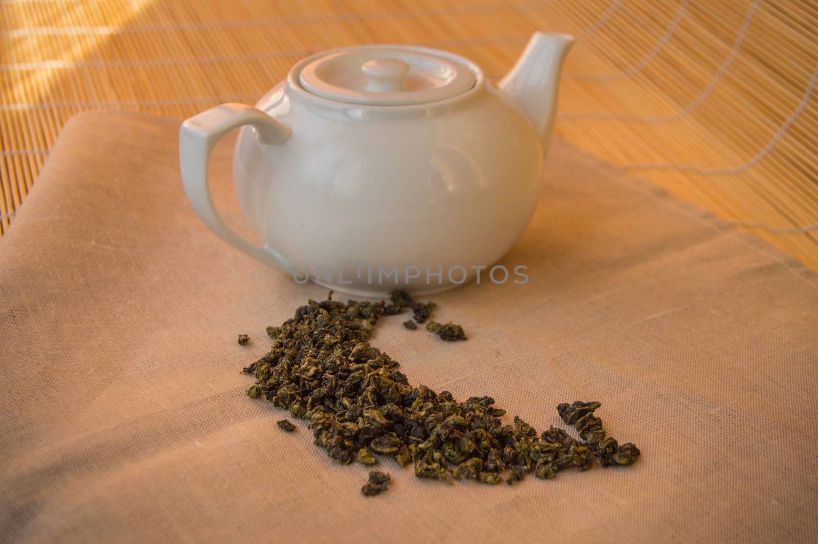 Teapot with green tea leaves on the napkin, bamboo background.