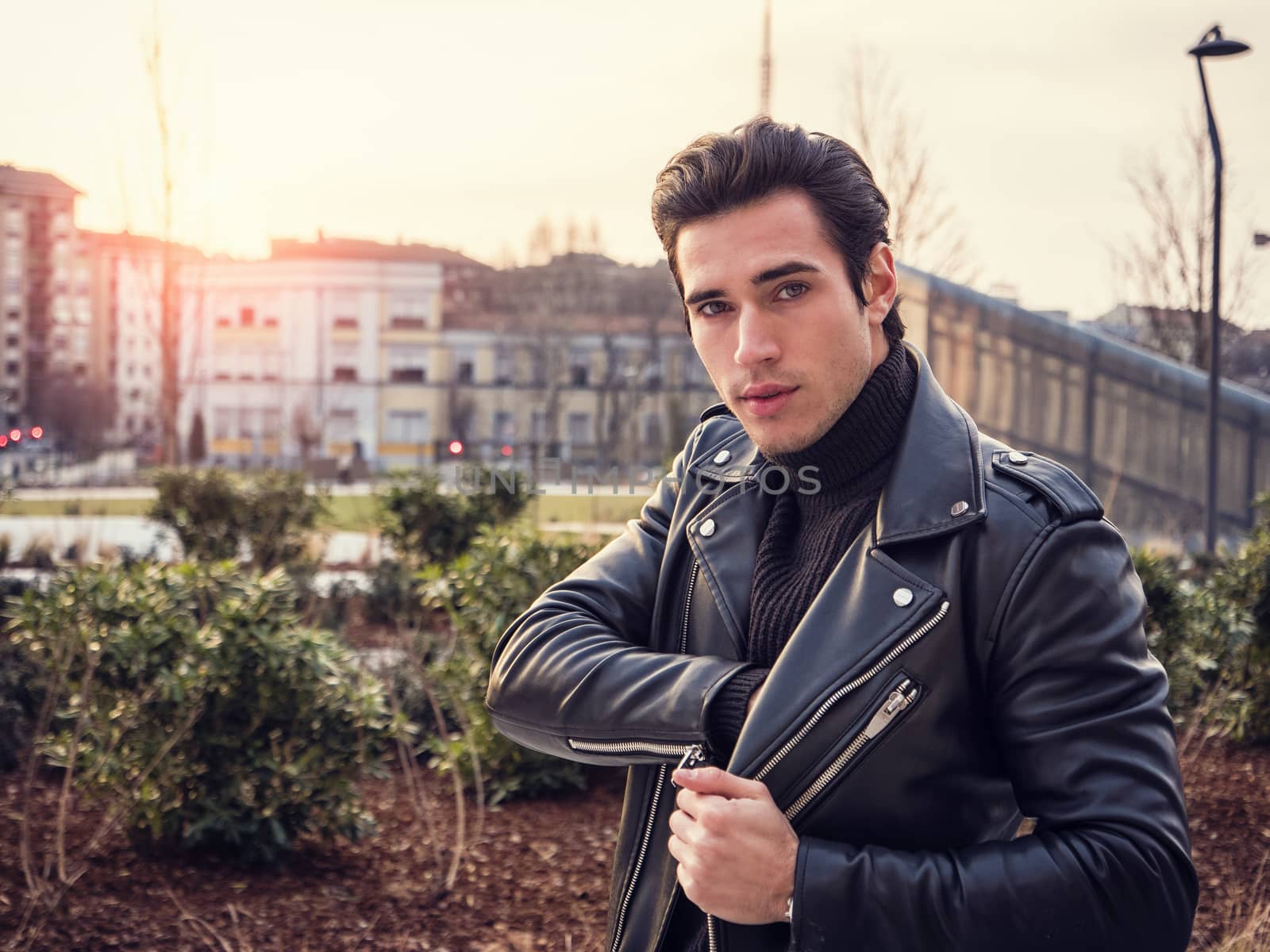 One handsome young man in urban setting in modern city, standing, wearing black leather jacket and jeans, looking at camera, at sunset