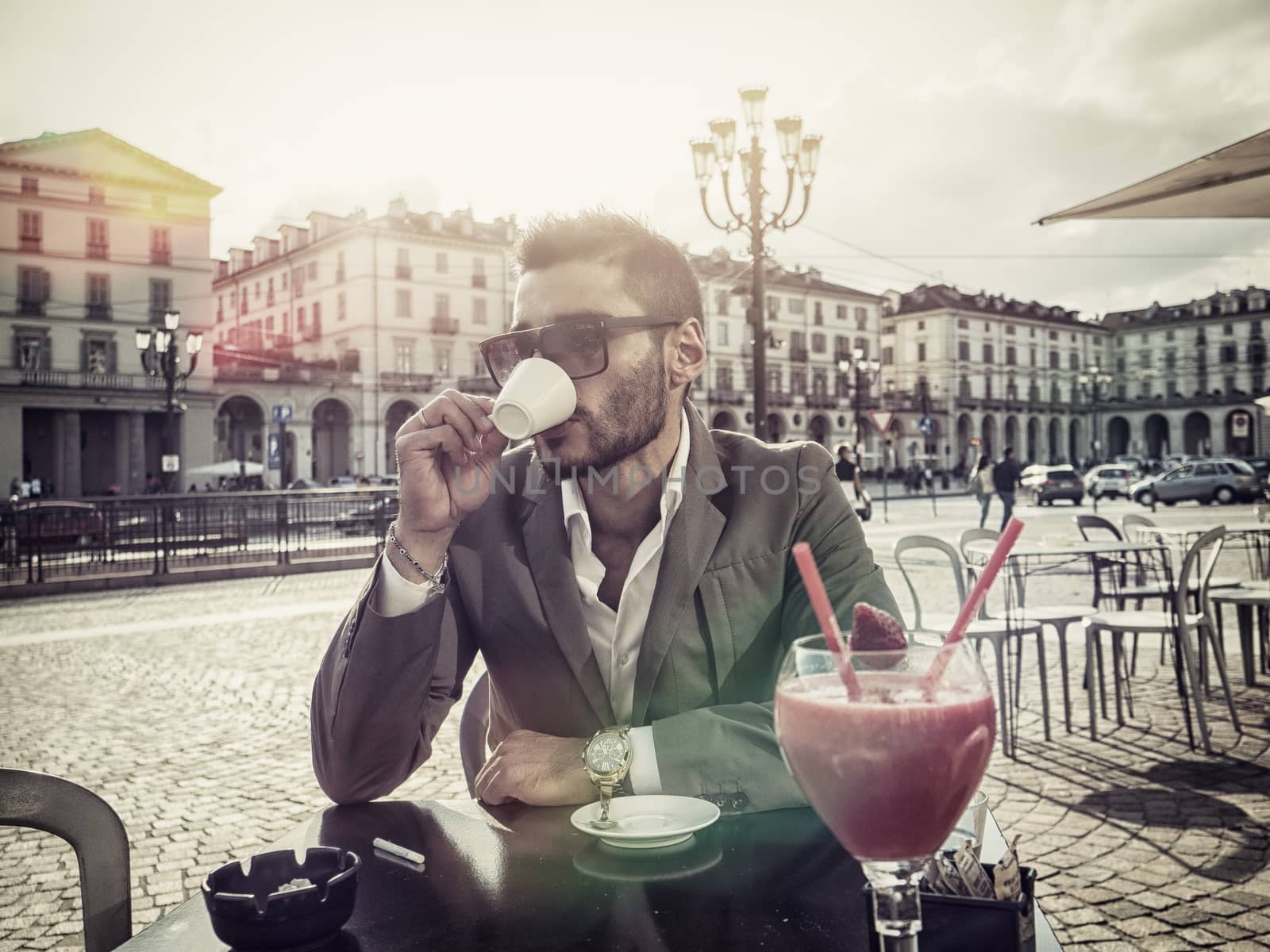 Handsome young man drinking espresso coffee, wearing elegant coat posing at table outside on urban background.