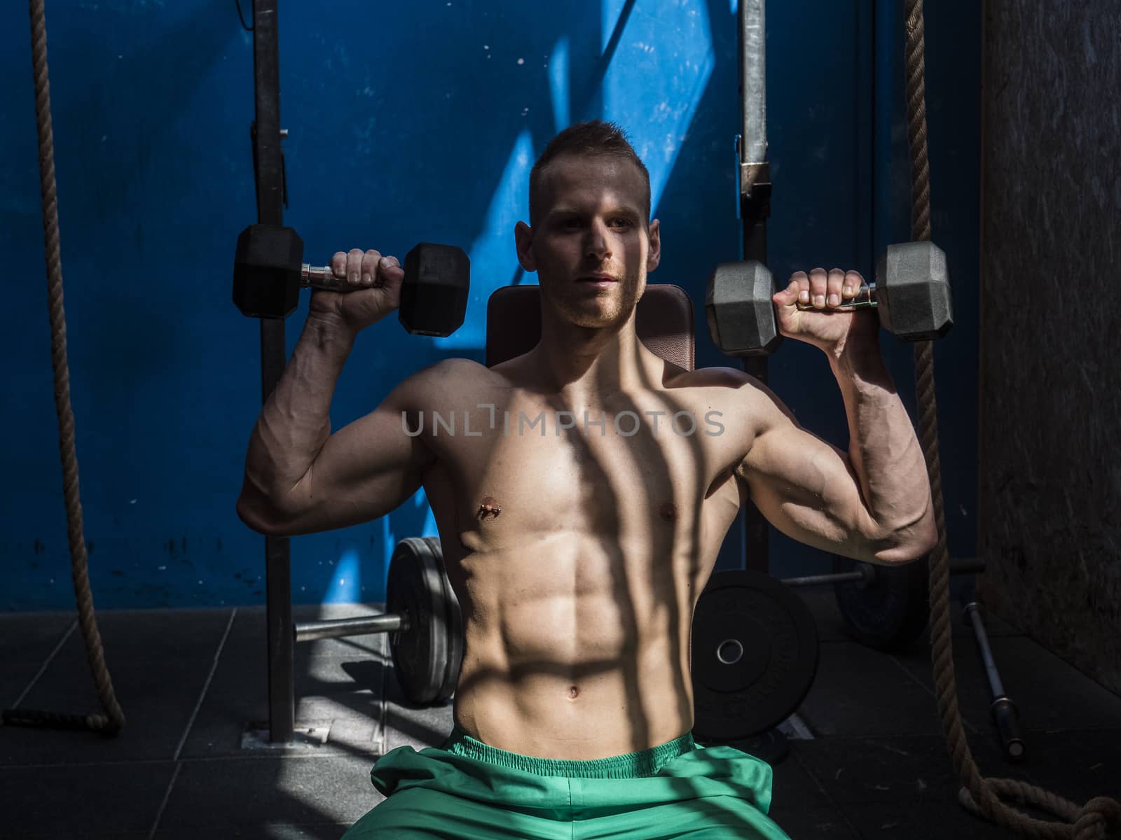 Attractive blond young man shirtless in gym working out, doing exercises for pecs.
