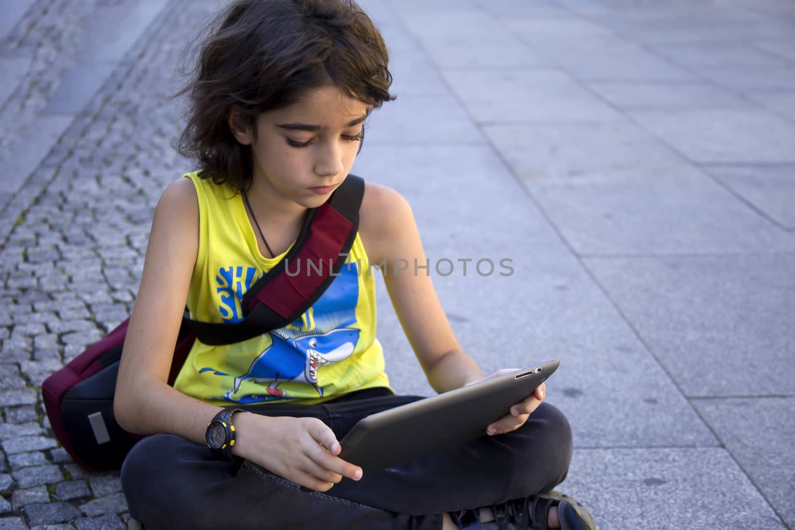 Teenager sitting outside on concrete using a tablet computer, looking down while sitting with crossed legs. by Anelik