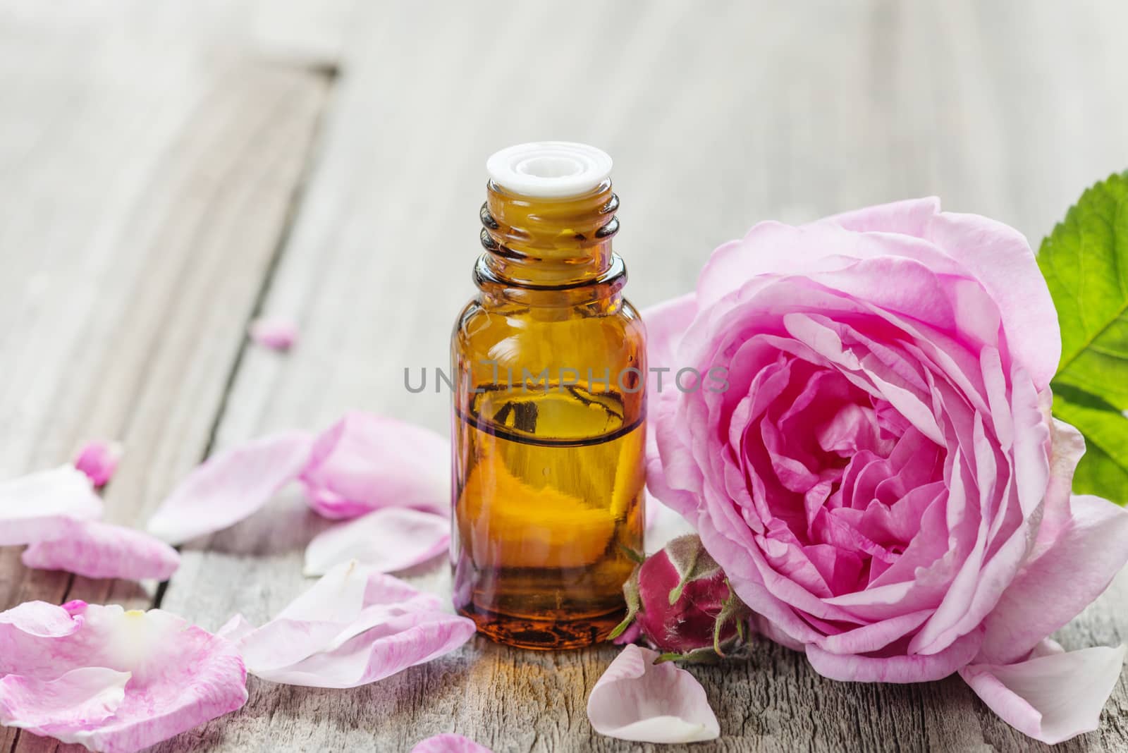 Dark glass vial with rose essential oil and flower of pink rose on a wooden background, with space for text
