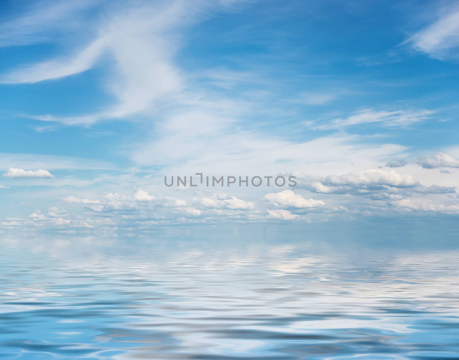 Panorama of vast blue summer sky with fluffy white cumulus and cirrus clouds reflected in a water surface with small waves