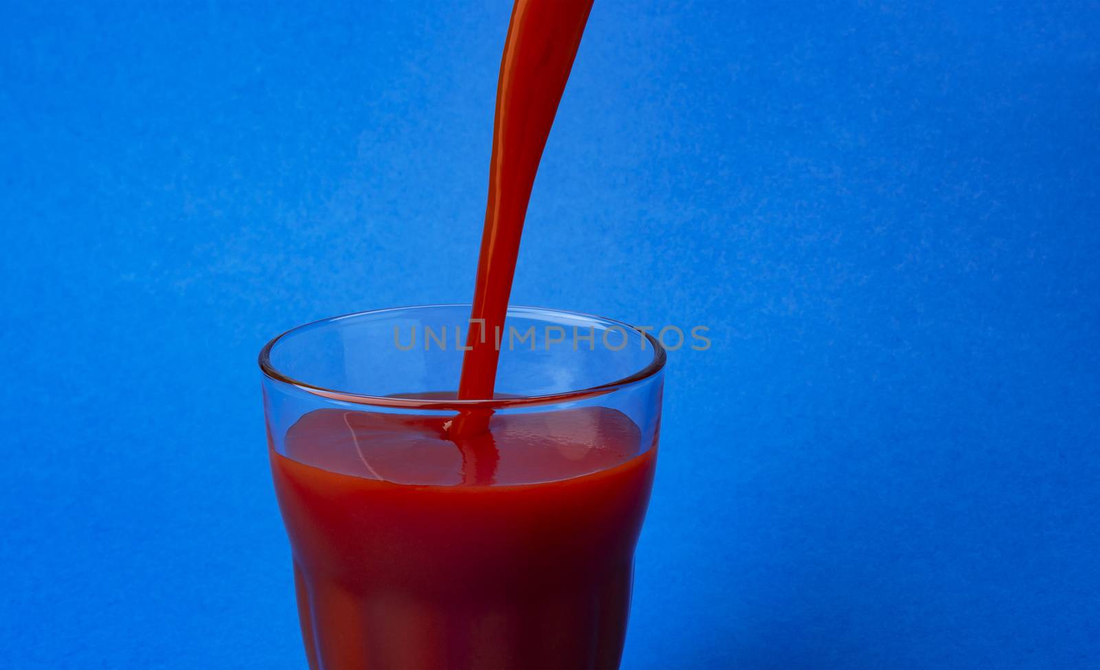 Tomato juice pouring into glass, isolated on blue background, with copy space, healthy drink concept, close-up