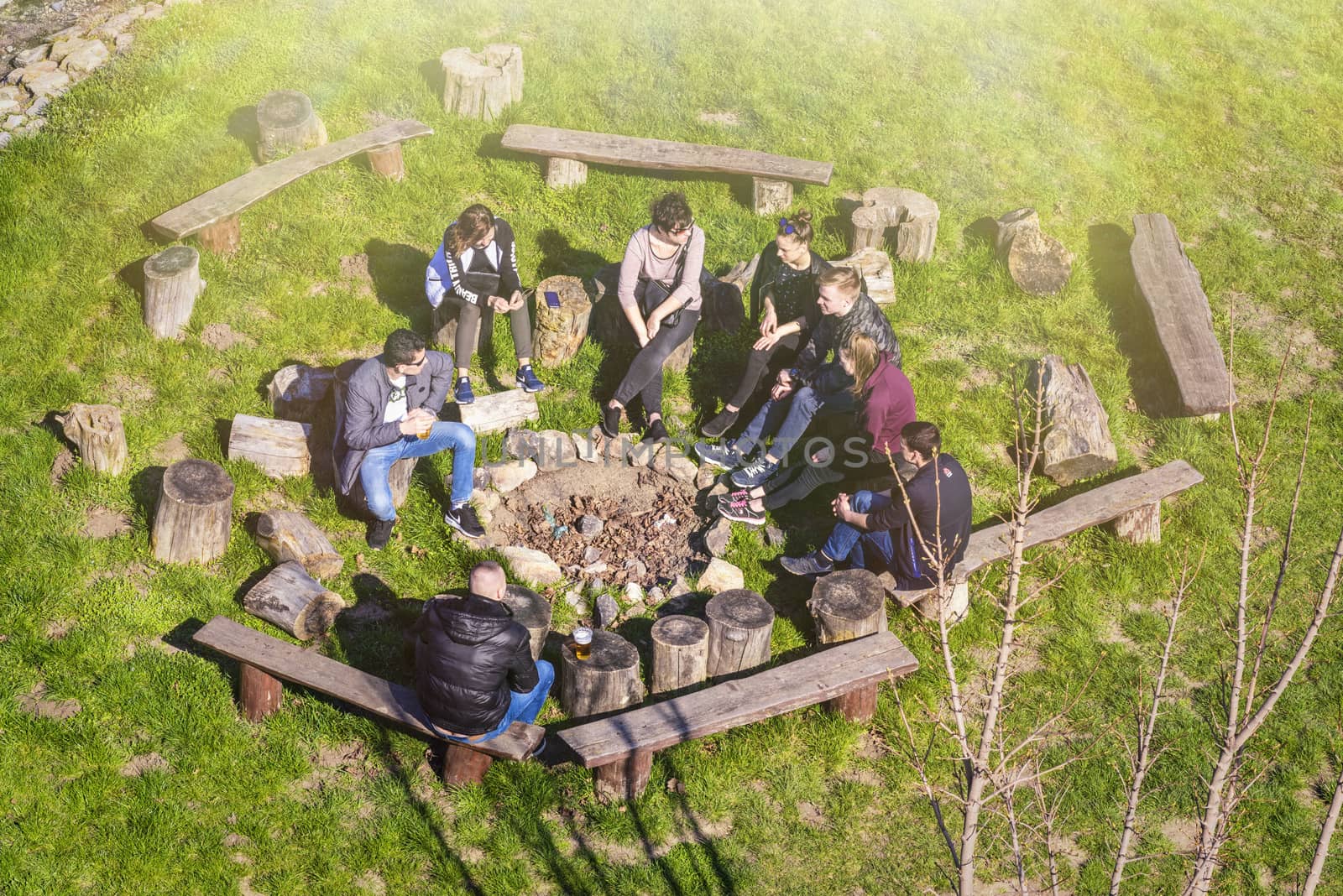A group of people are sitting by the fire. in a camp with a guitar, tent, blanket, backpacks, sitting near the fire place in a camp.Spring warm day.