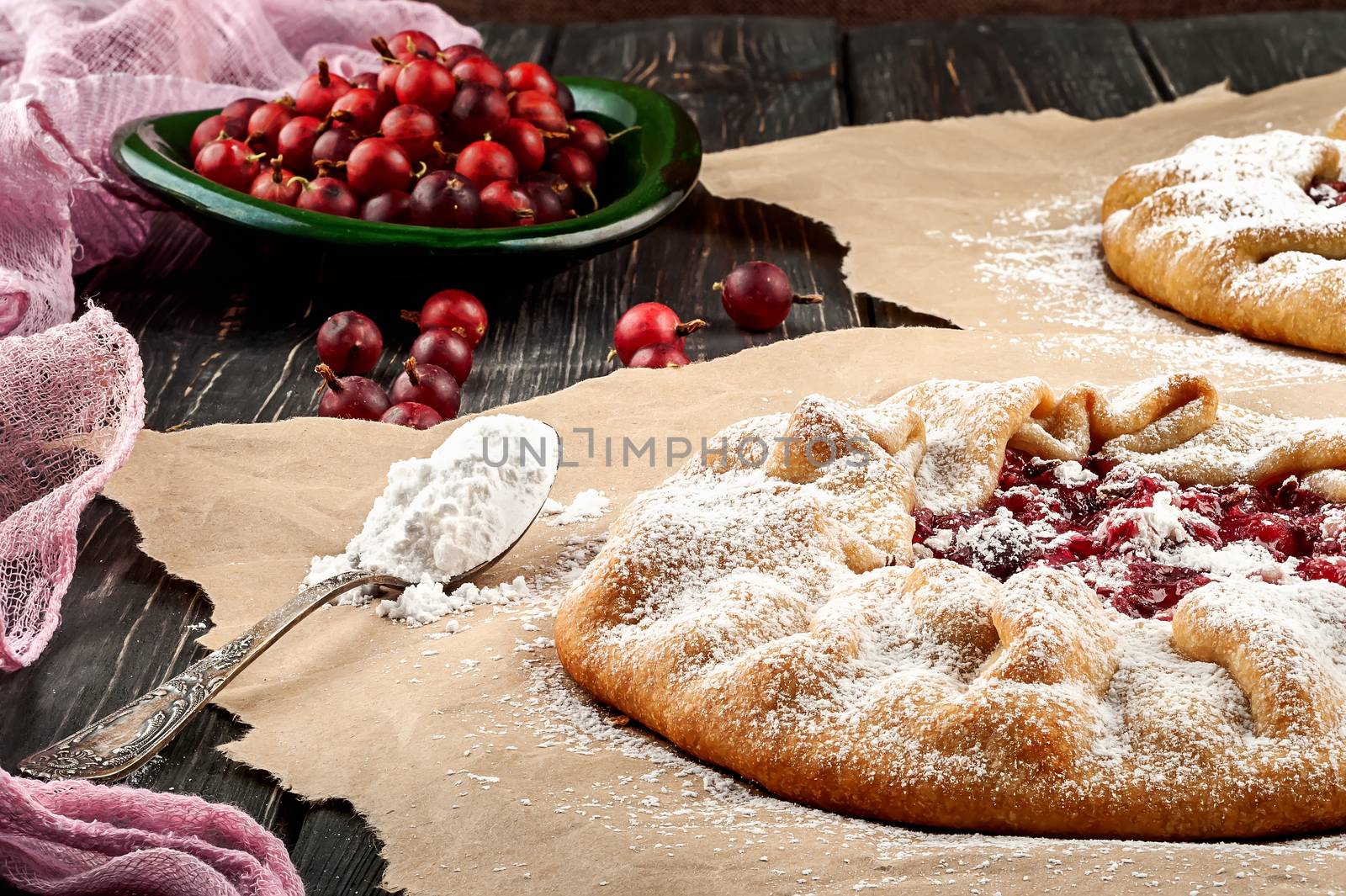 Open pies with gooseberries on a wooden table. Spoon with powdered sugar nearby. Gooseberry on a green plate in the background.