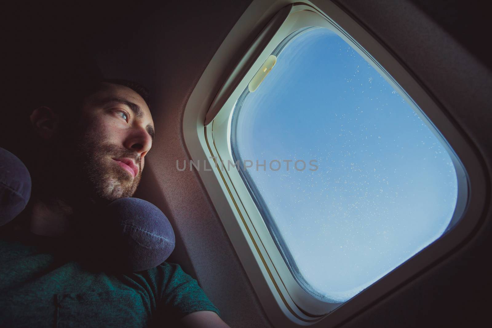 Pensive man with neck pillow looking outside through the window of an airplane.