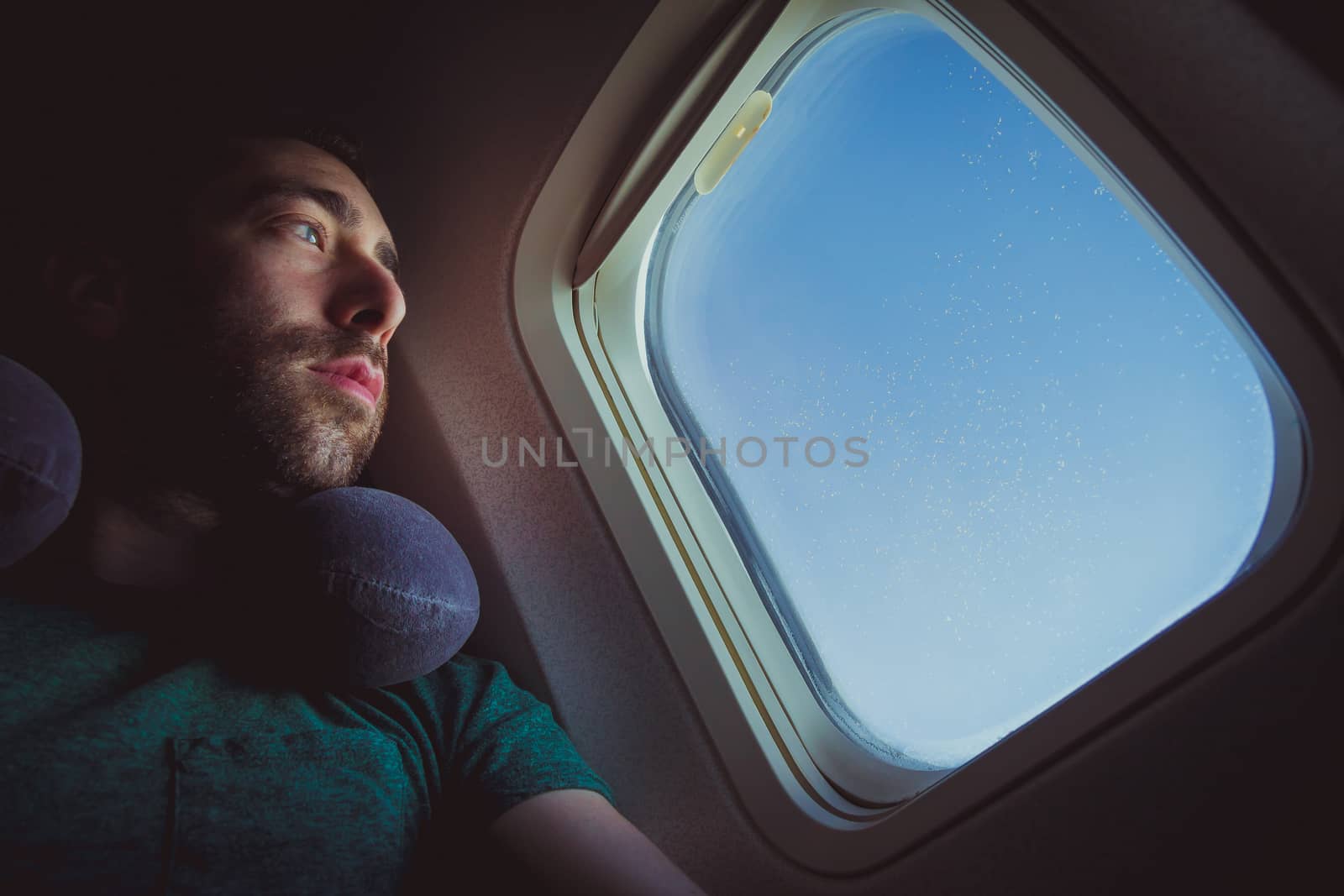 Pensive man with neck pillow looking outside through the window of an airplane.
