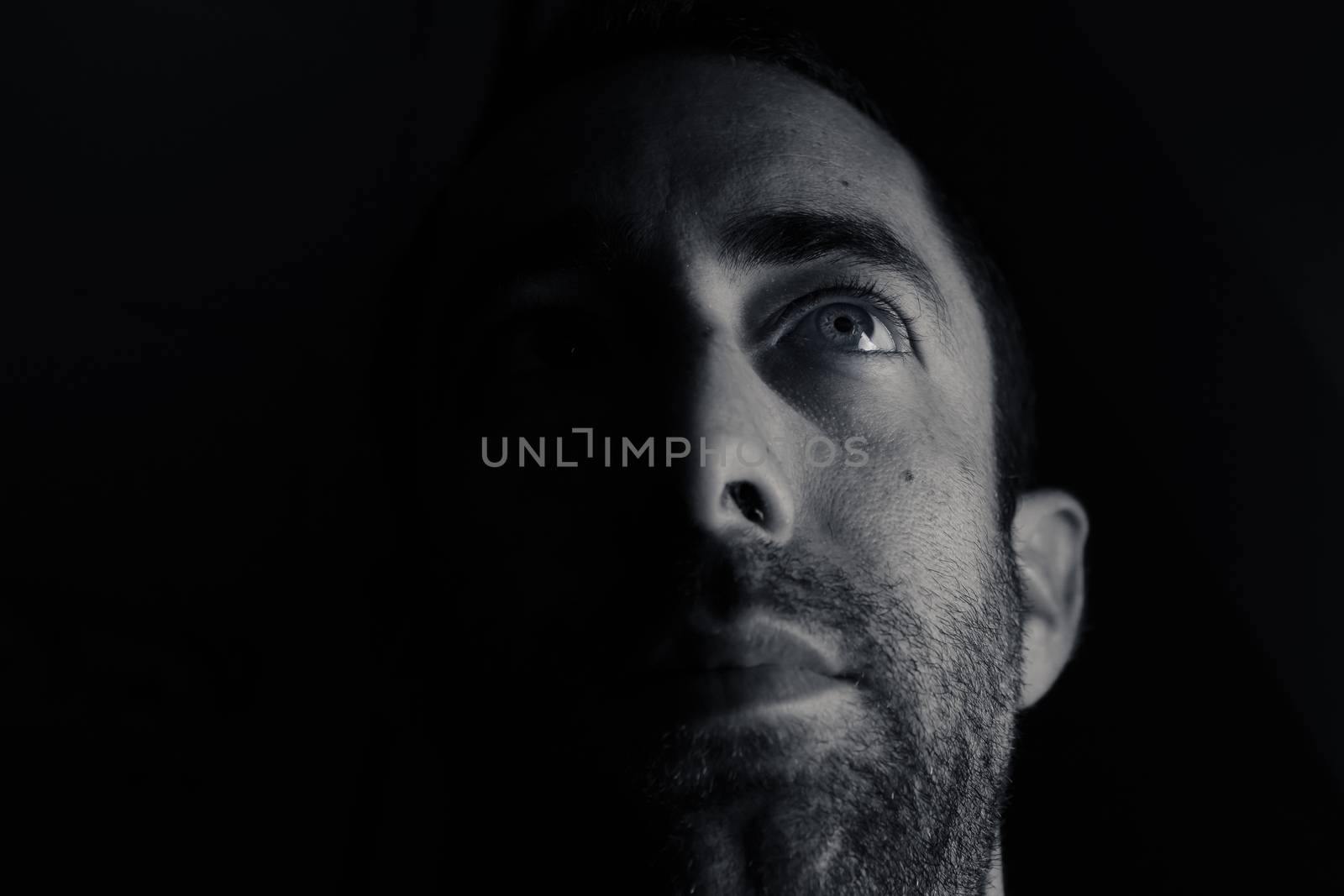 Dramatic portrait of a young man half expressionless face in deep shadow looking up isolated on black background.