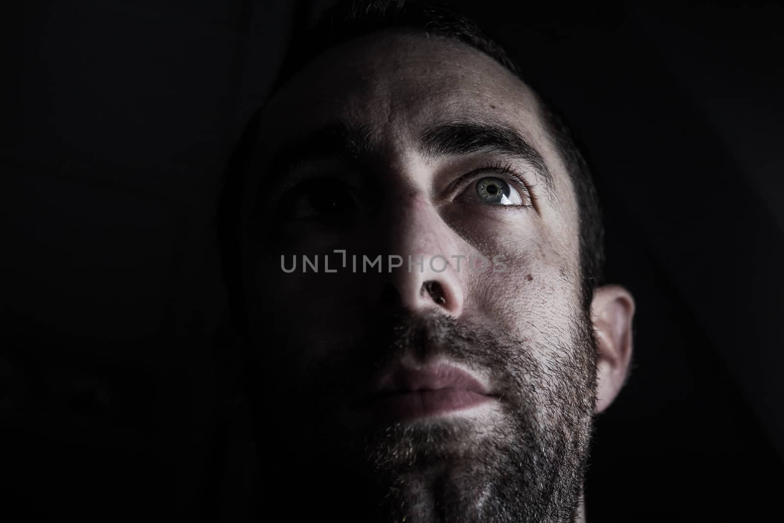 Dramatic portrait of a young man half expressionless face in deep shadow looking up isolated on black background.