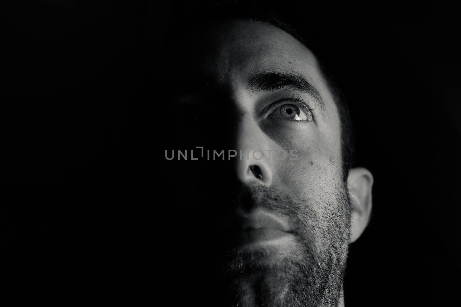 Dramatic portrait of a young man half expressionless face in deep shadow looking up isolated on black background.