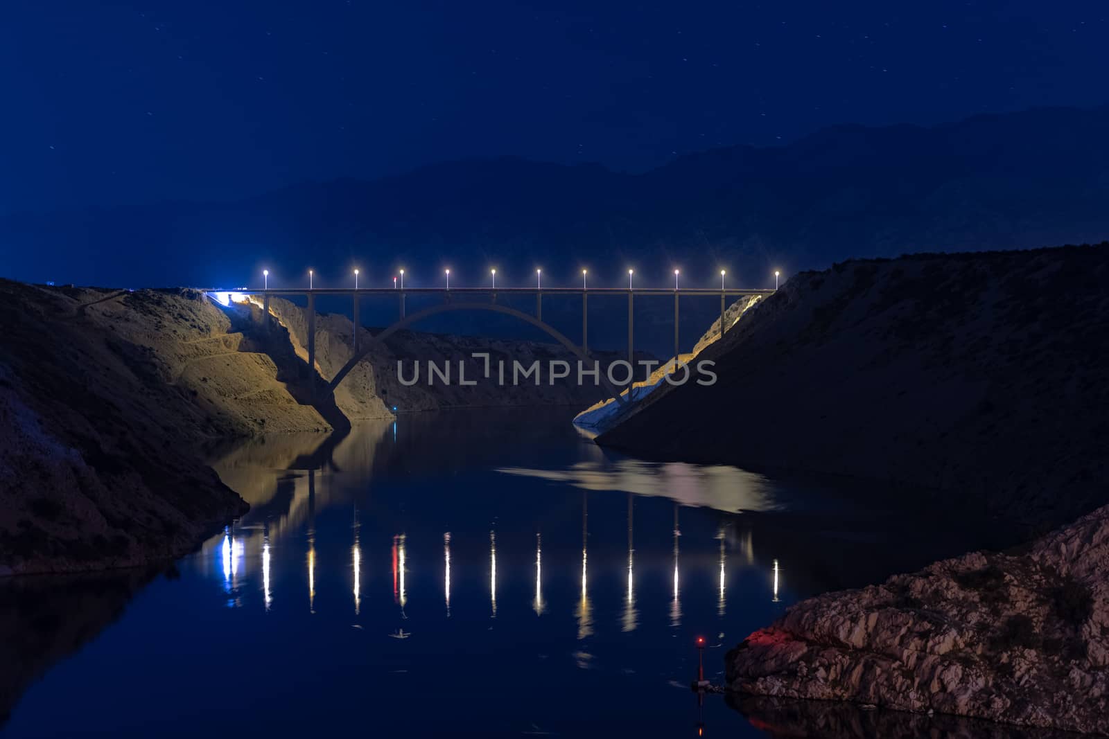 Illuminated highway bridge under stars, reflection of lights in water, mountains in background and bright stars in sky by asafaric