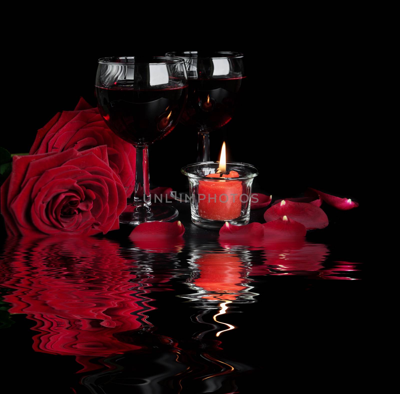 Valentine's Days concept: two glasses of red wine, two red roses and a burning red candle isolated on a black background reflected in a water surface with small waves