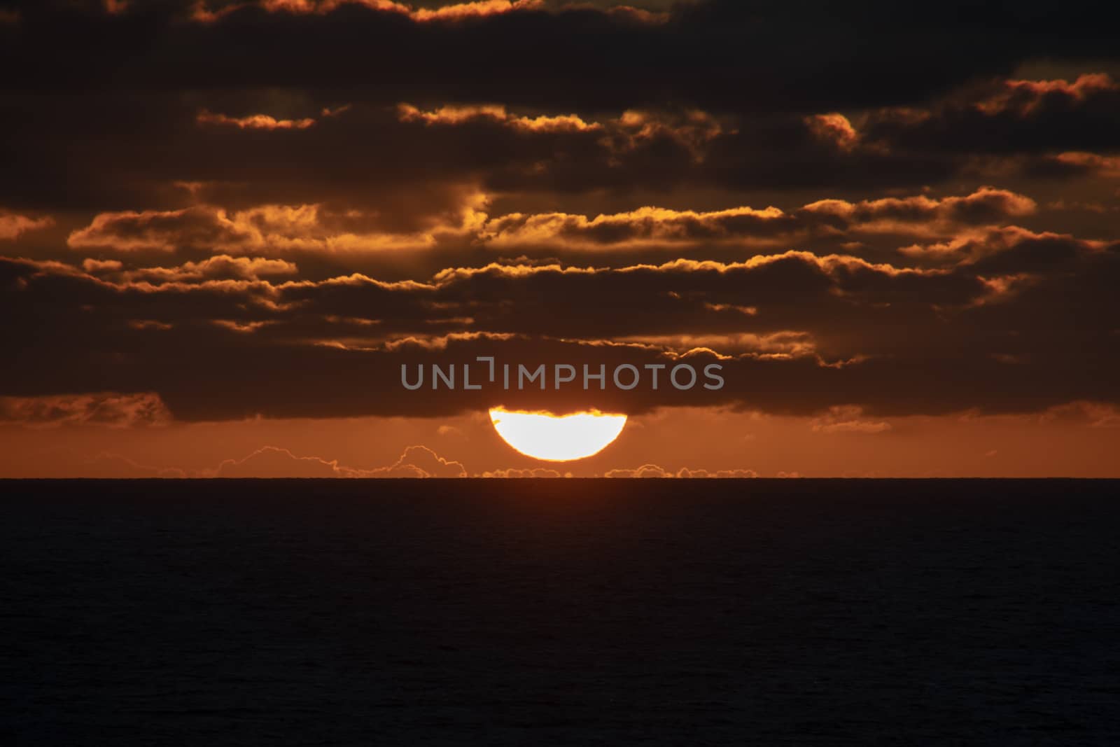 Sun a short moment before touching the horizon partly covered by red shining clouds by MXW_Stock