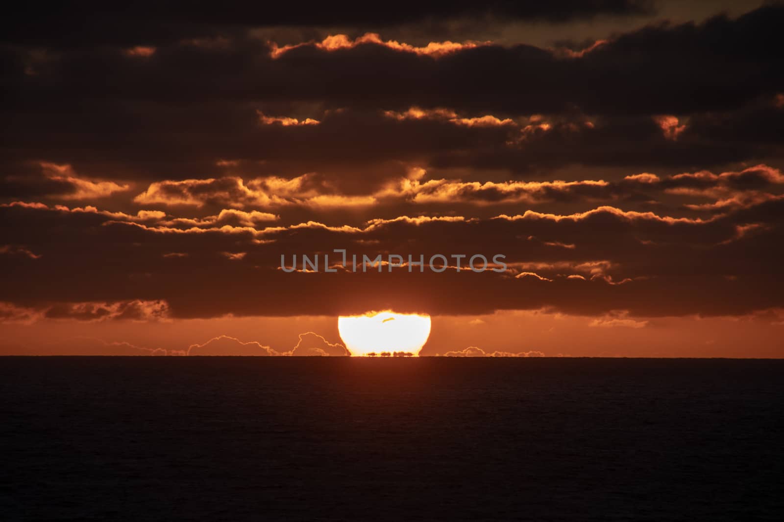 Sun party set under the horizon and partly covered by red shining clouds during sunset in Geraldton, Australia by MXW_Stock