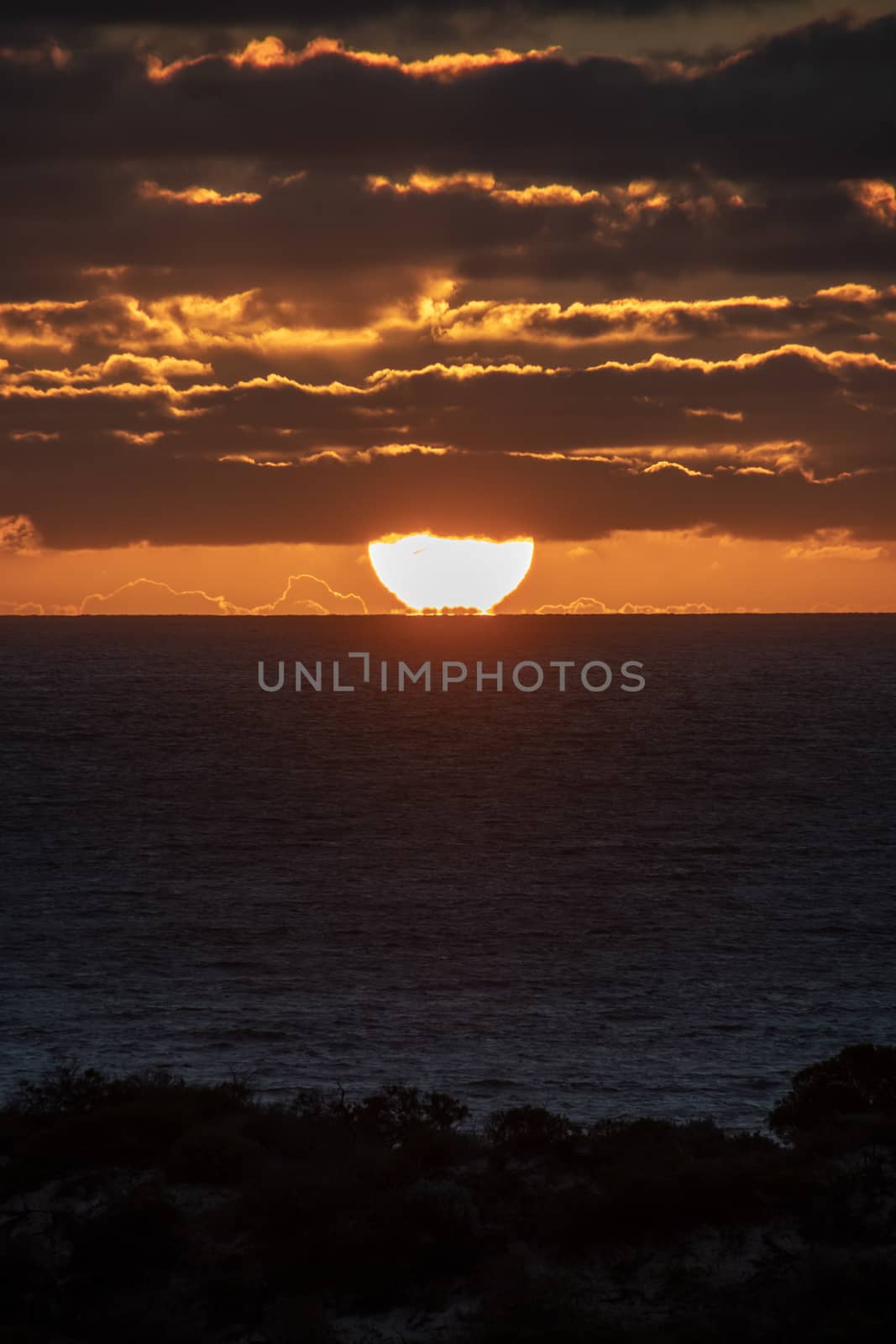 Sun just touching the horizon partly covered by red shining clouds during sunset in Geraldton, Australia by MXW_Stock