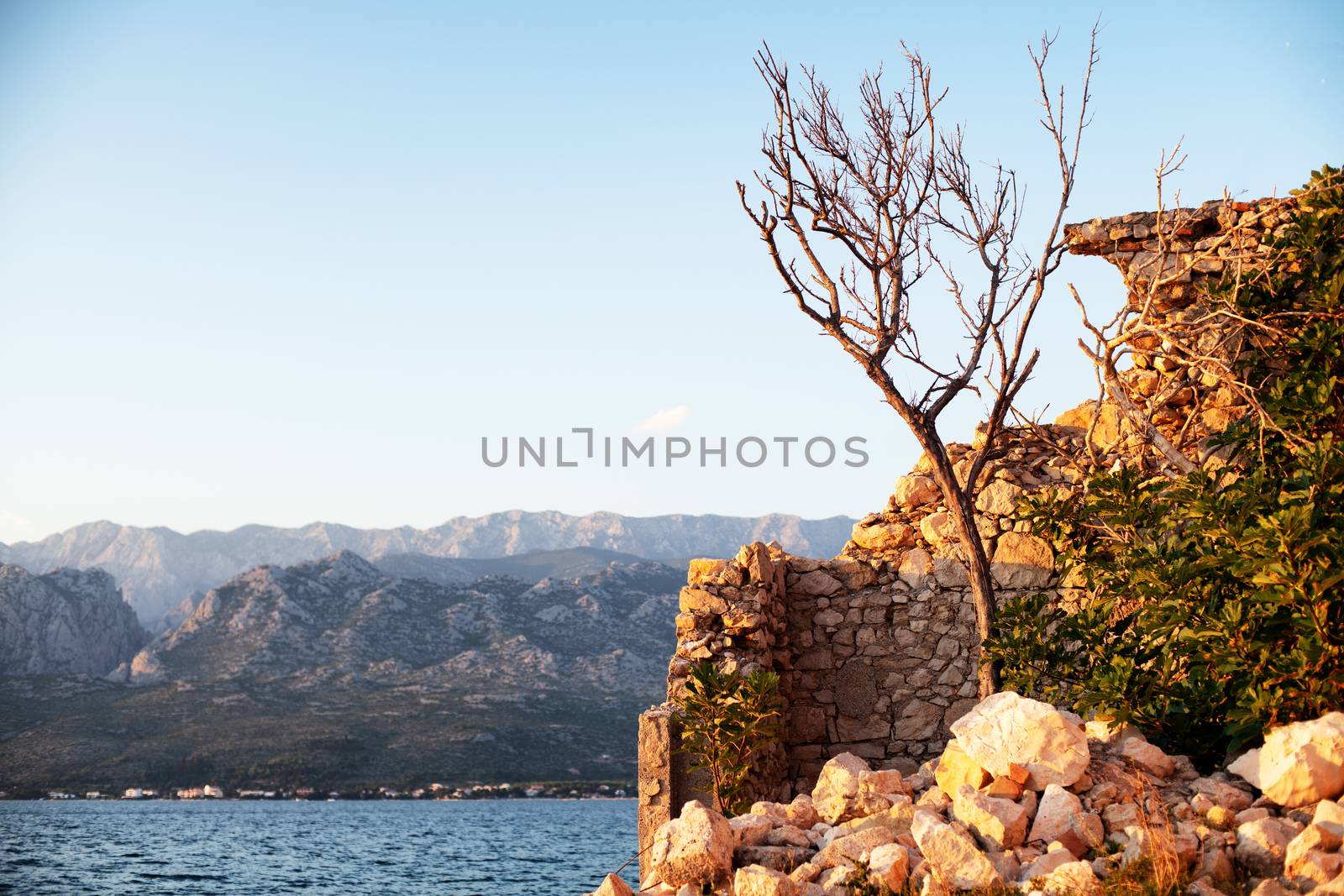 Ruins of stone house on waterfront in sunset, mountains and sea in background by asafaric