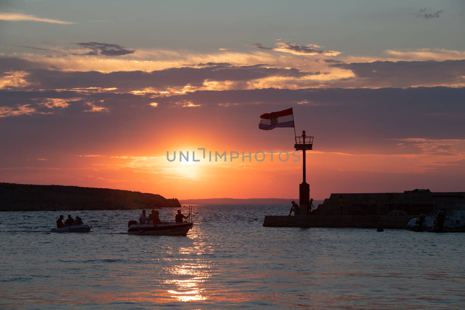 Croatian flag flying in wind at sunset in harbor, all people silhuetted and unrecognizable, small boats in front entering harbor