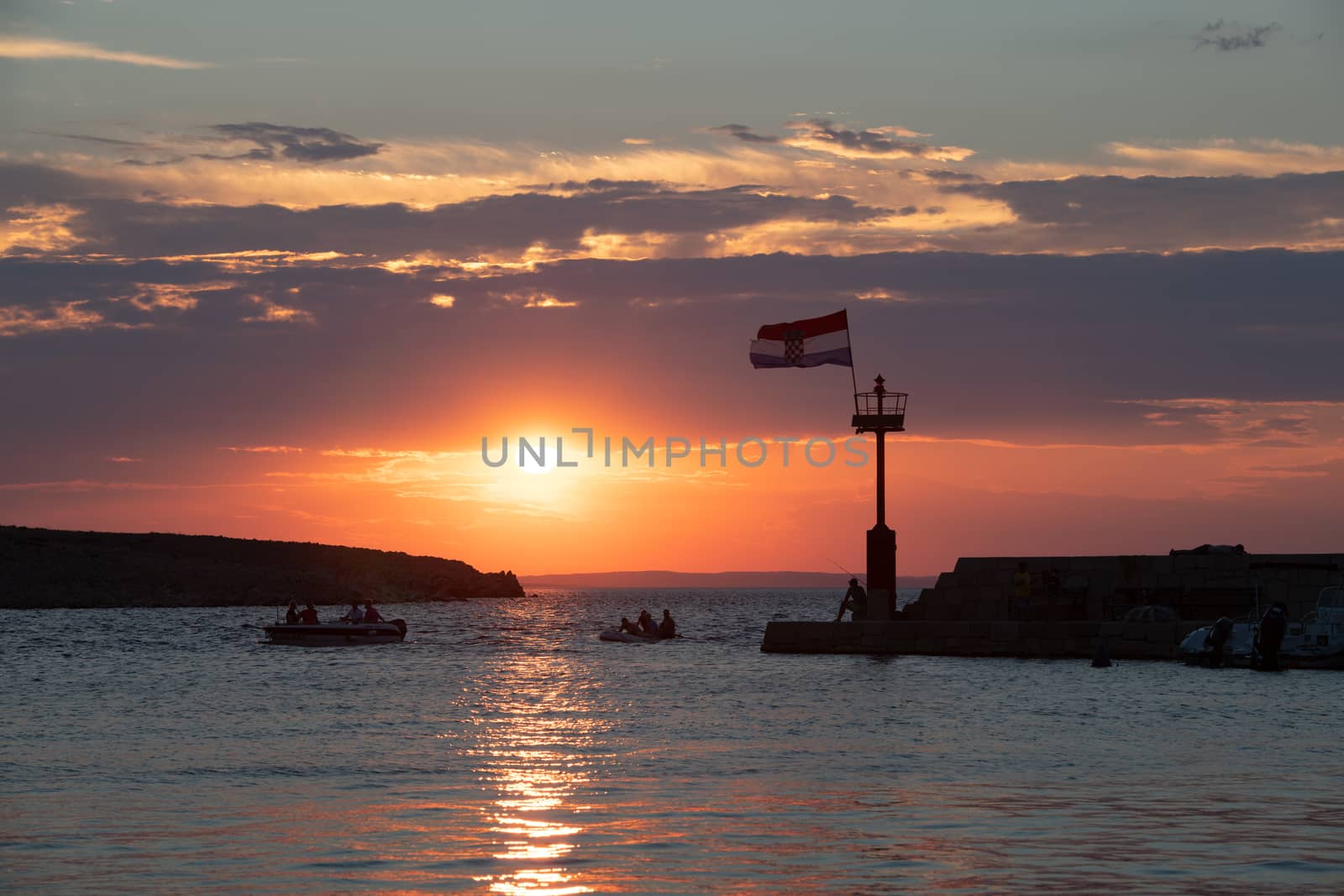 Croatian flag flying in wind at sunset in harbor, all people silhuetted and unrecognizable, small boats in front near lighthouse