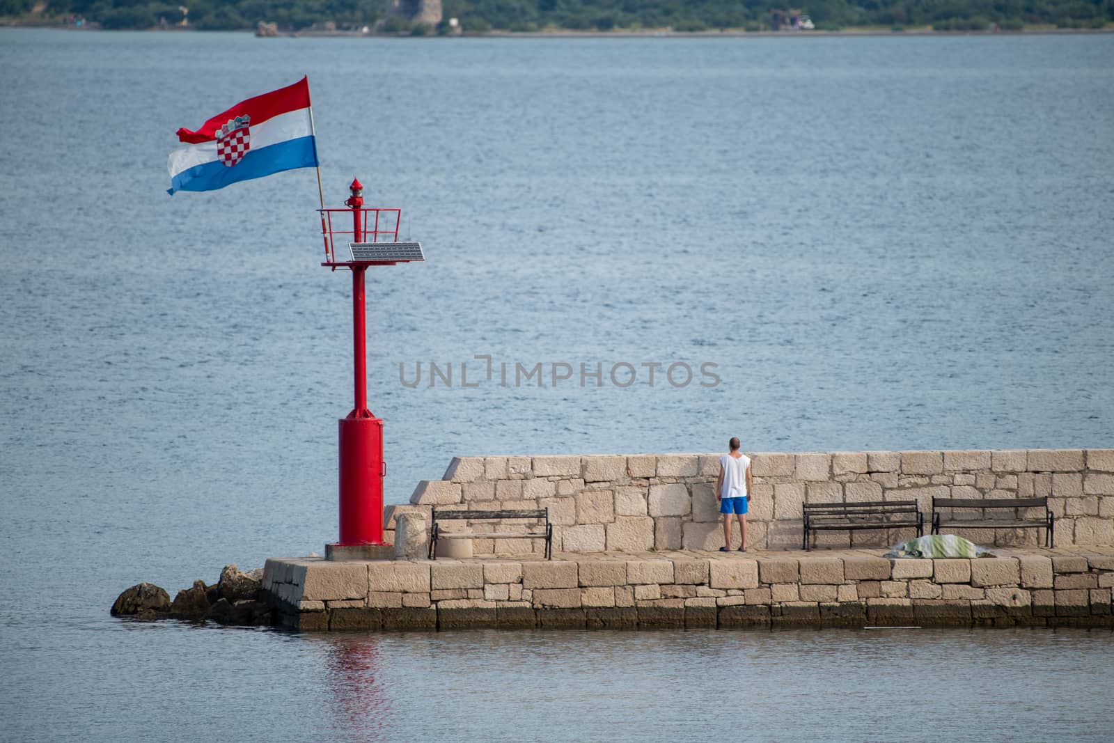 Croatian flag flying in wind on lighthouse in harbor, daytime, stone pier, man, facing away looking over stone wall by asafaric