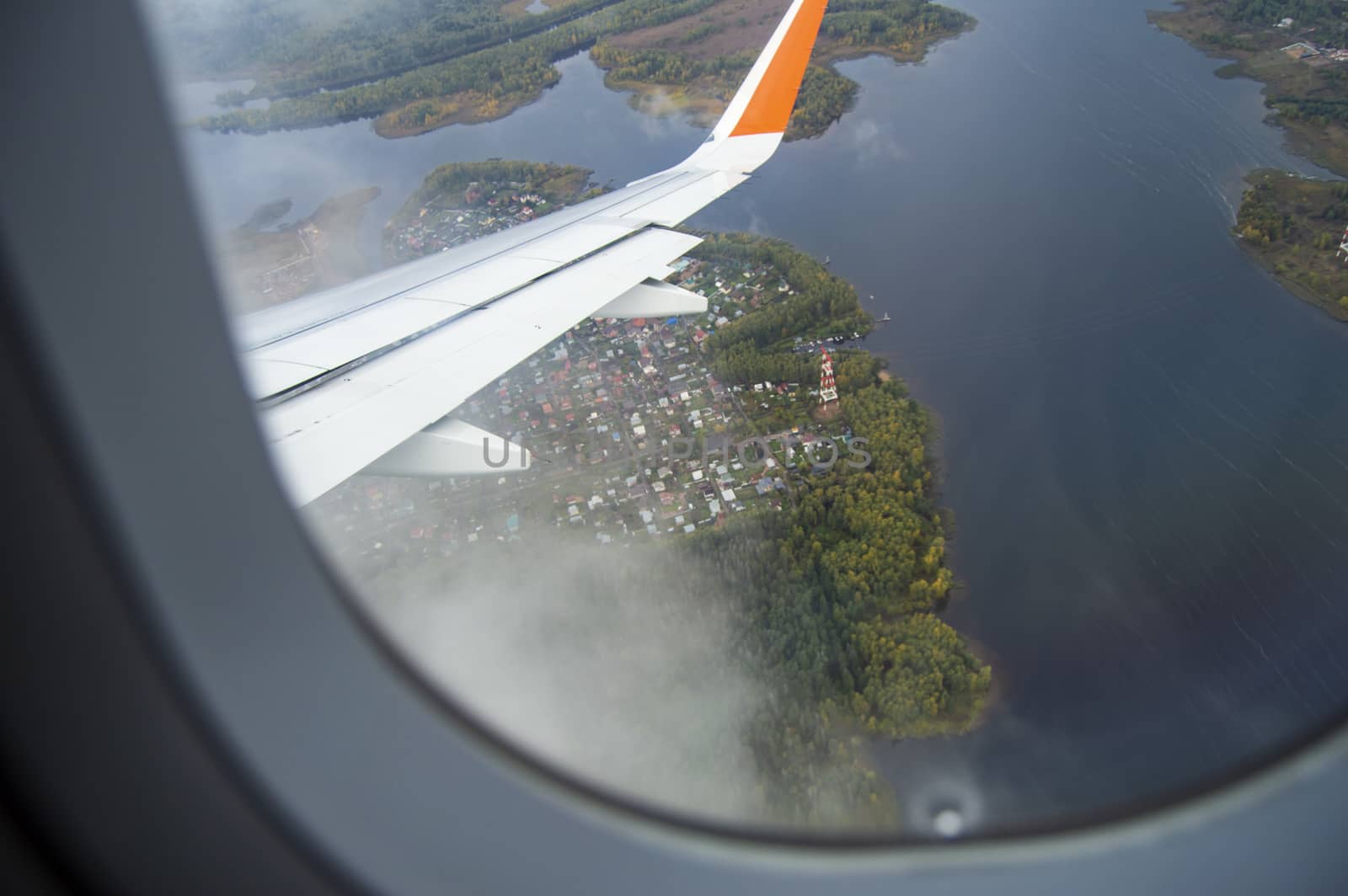 View from the window of the aircraft on the buildings and trees, the wing of the aircraft on the background of passing clouds, the concept of flight by claire_lucia