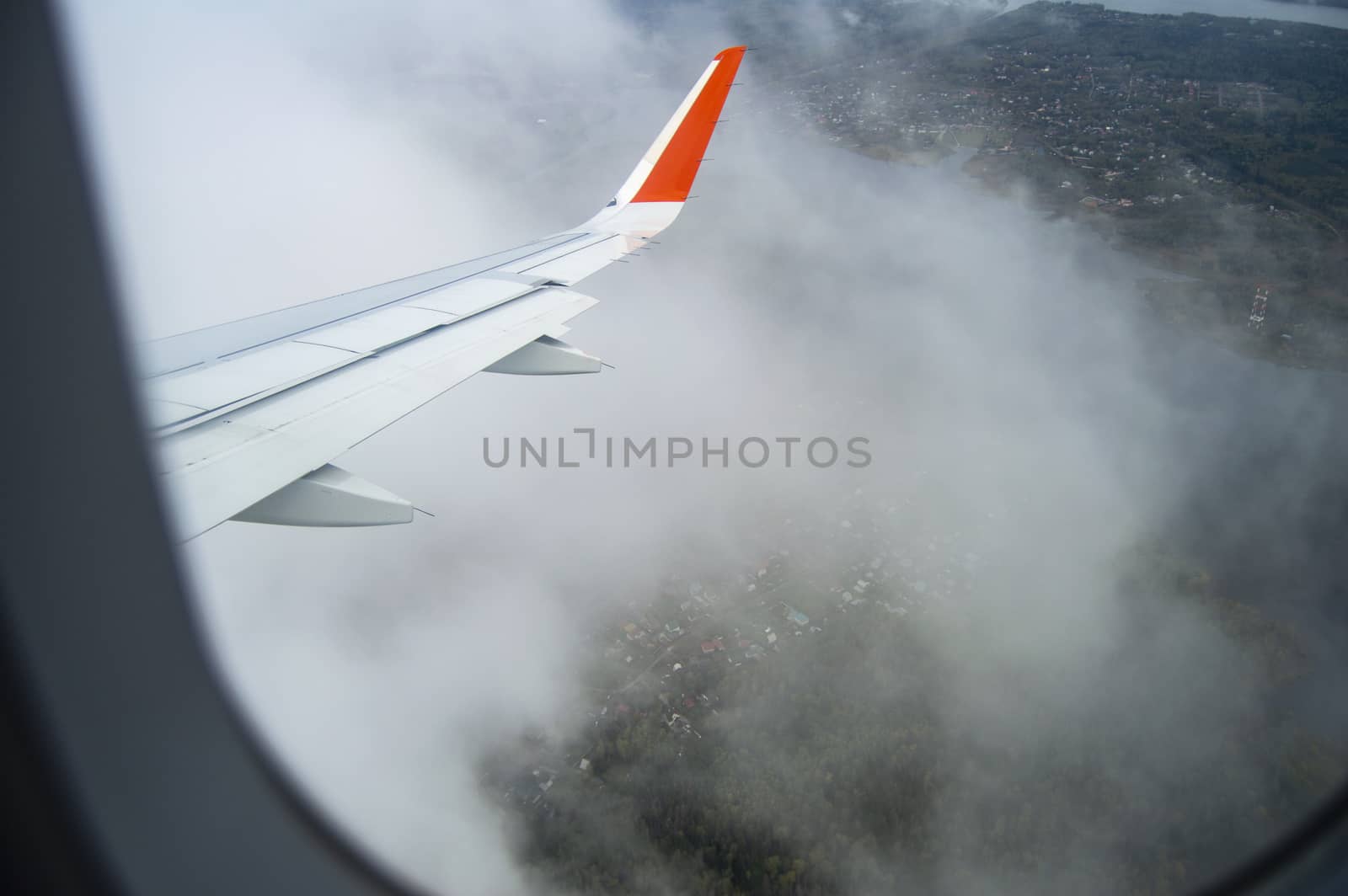 View from the window of the aircraft on the buildings and trees, the wing of the aircraft on the background of passing clouds, the concept of flight by claire_lucia