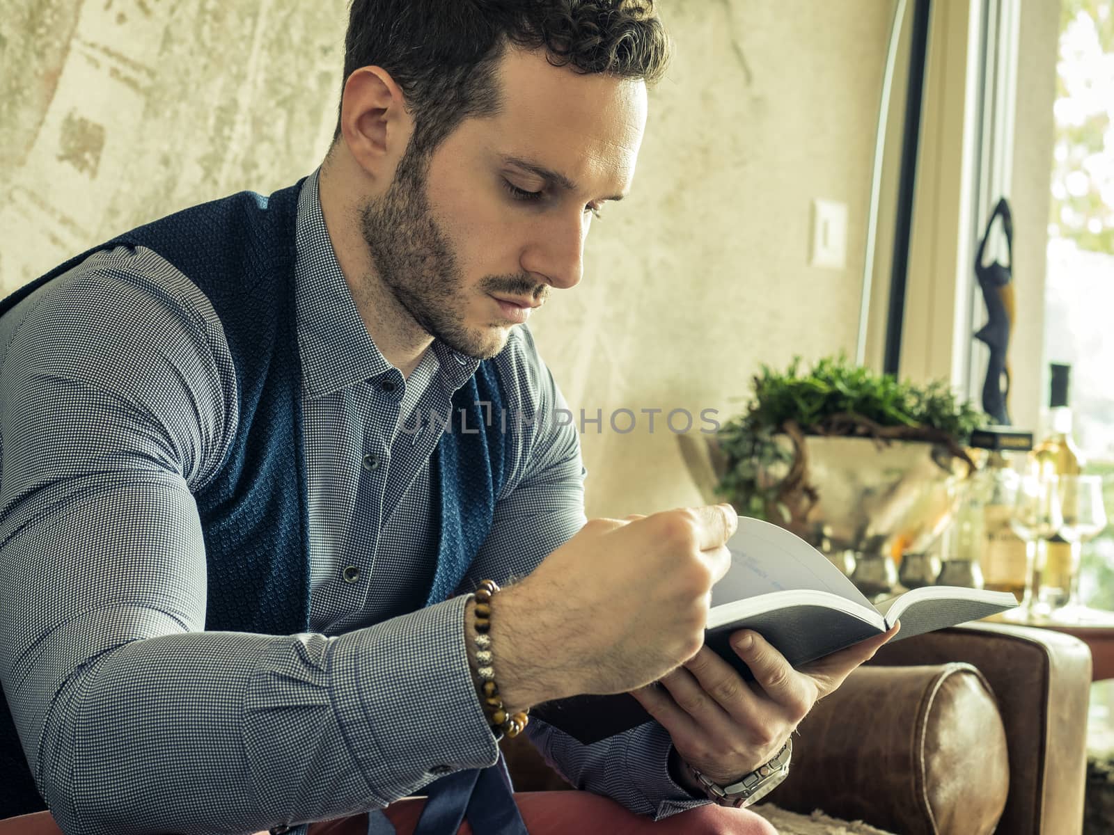 Handsome young man reading book at home in his living-room, sitting on sofa