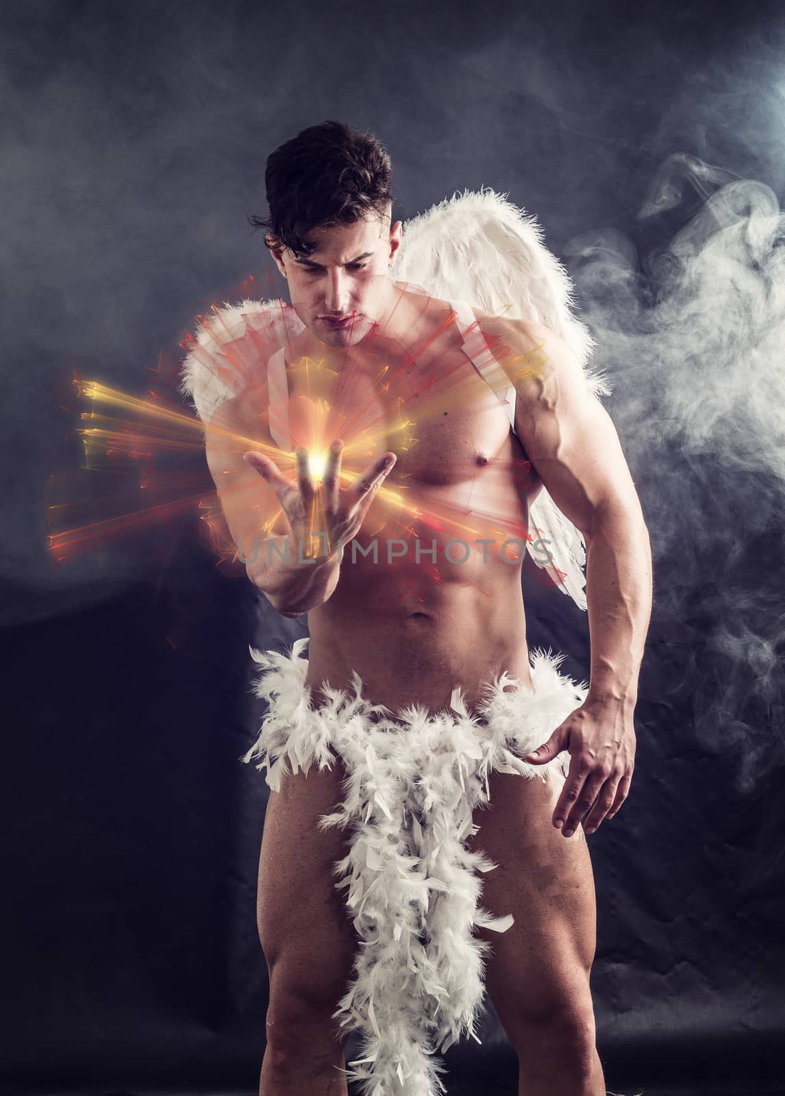 Naked male angel sitting in dark with white feather wings spreading from his back, holding ball of energy in hand. Angelic naked young man standing on dark background