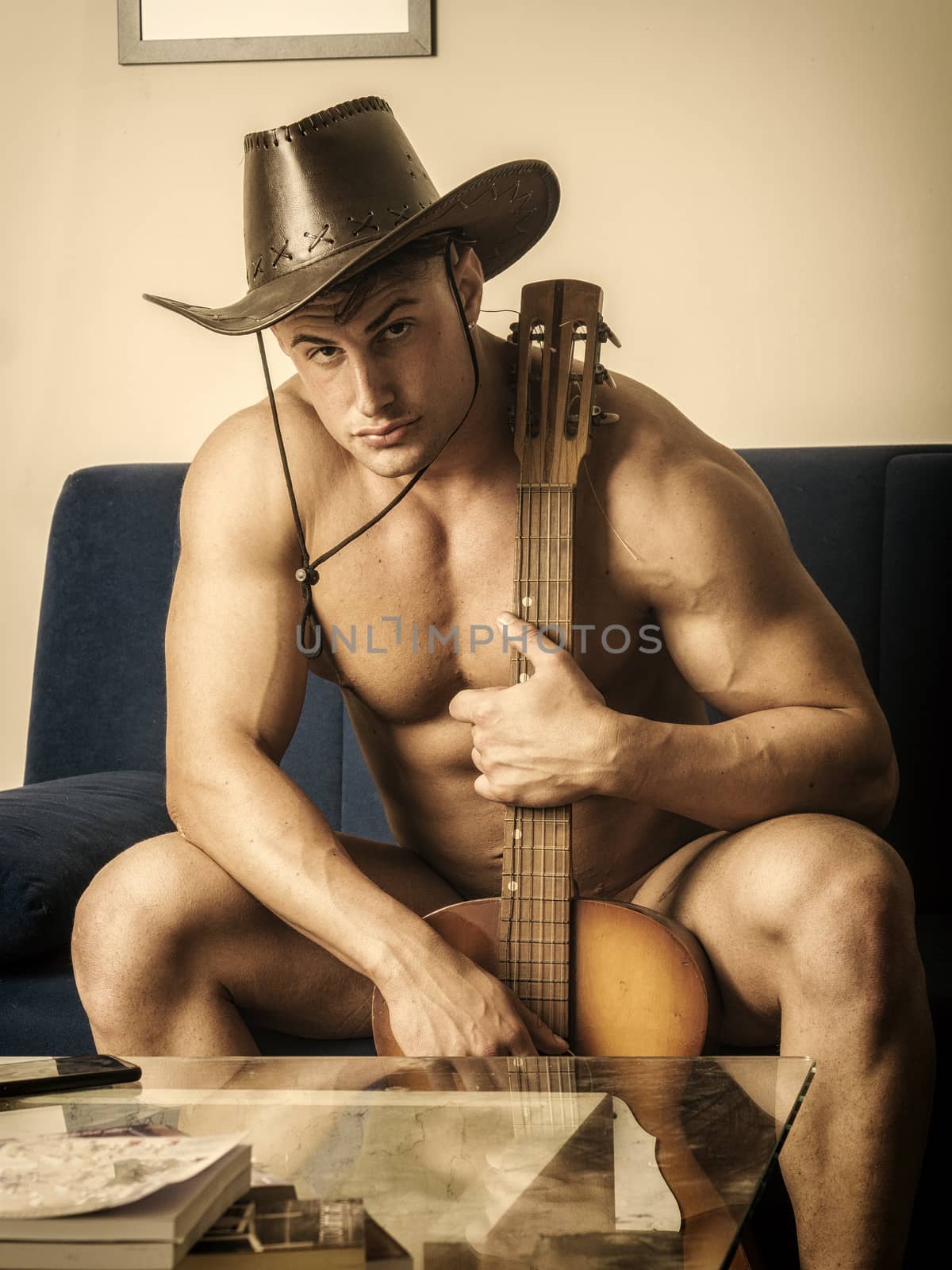 Portrait of handsome muscular young shirtless man playing guitar over wearing cowboy hat