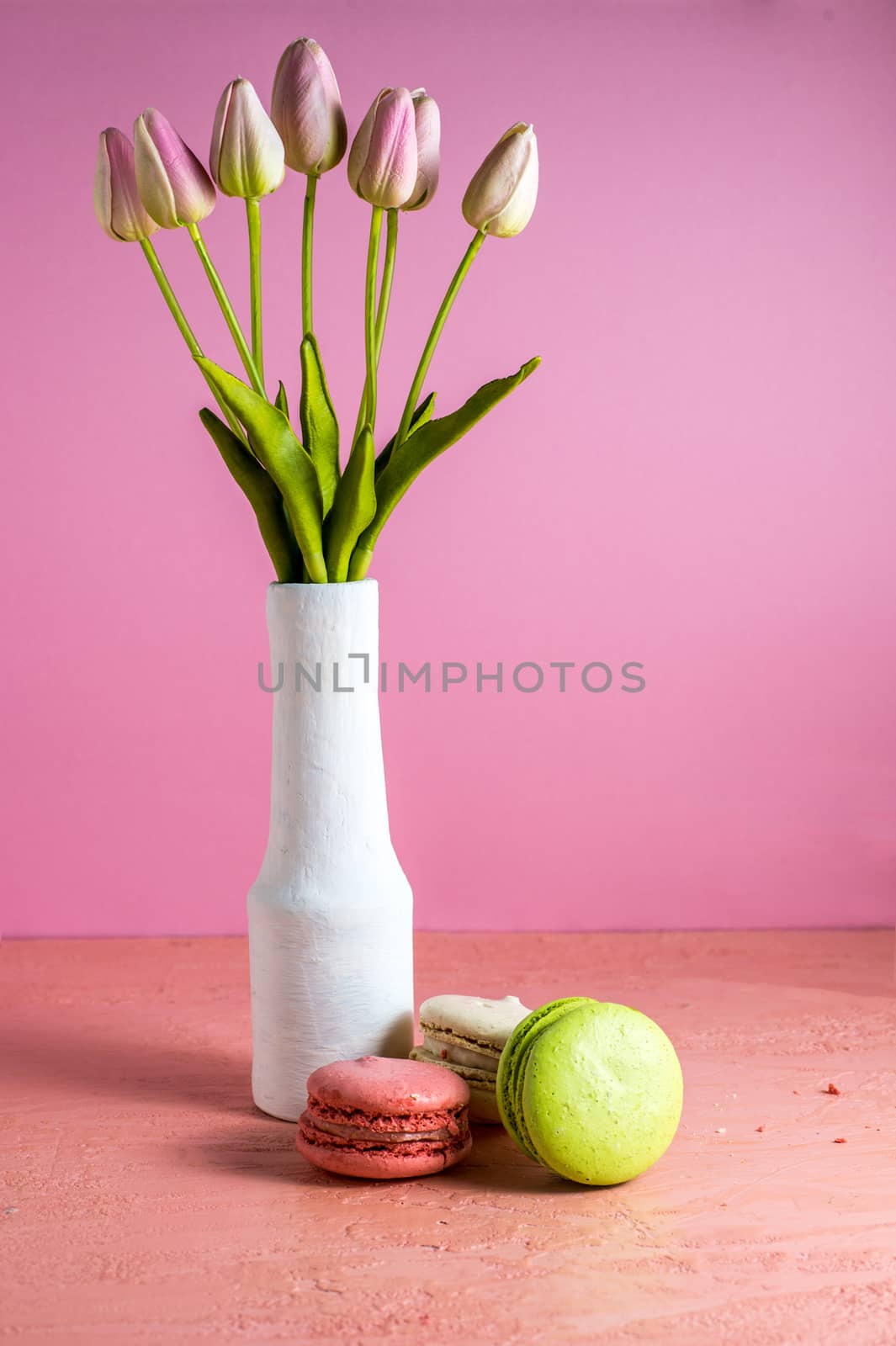 Makaroons on a pink background next to a bouquet of tulips in a white vase and copy space.