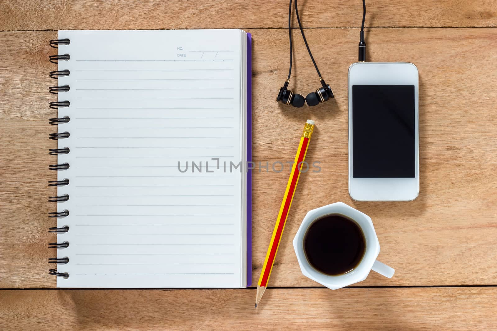 Bank notebook with pencil laying on the brown table. White mobile with earphones and black coffee put on the table as well.