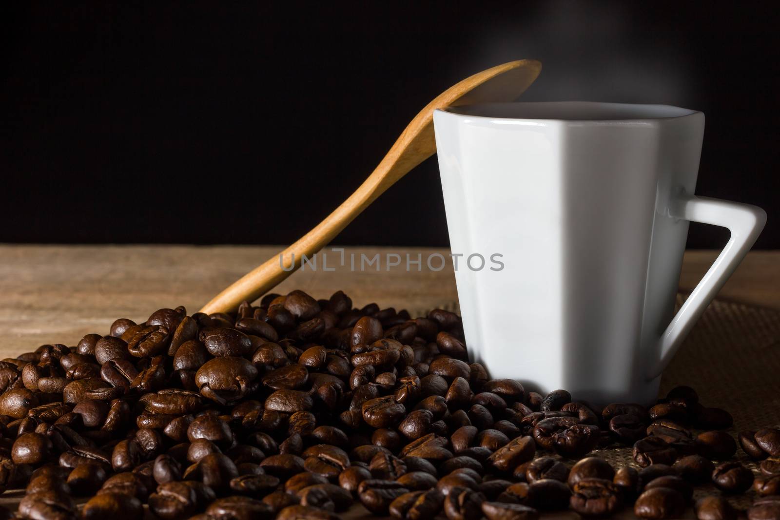 Low key coffee in the darkness. Roasted coffee beans on a wooden table with a glass of white ceramic and a wooden spoon in morning light.