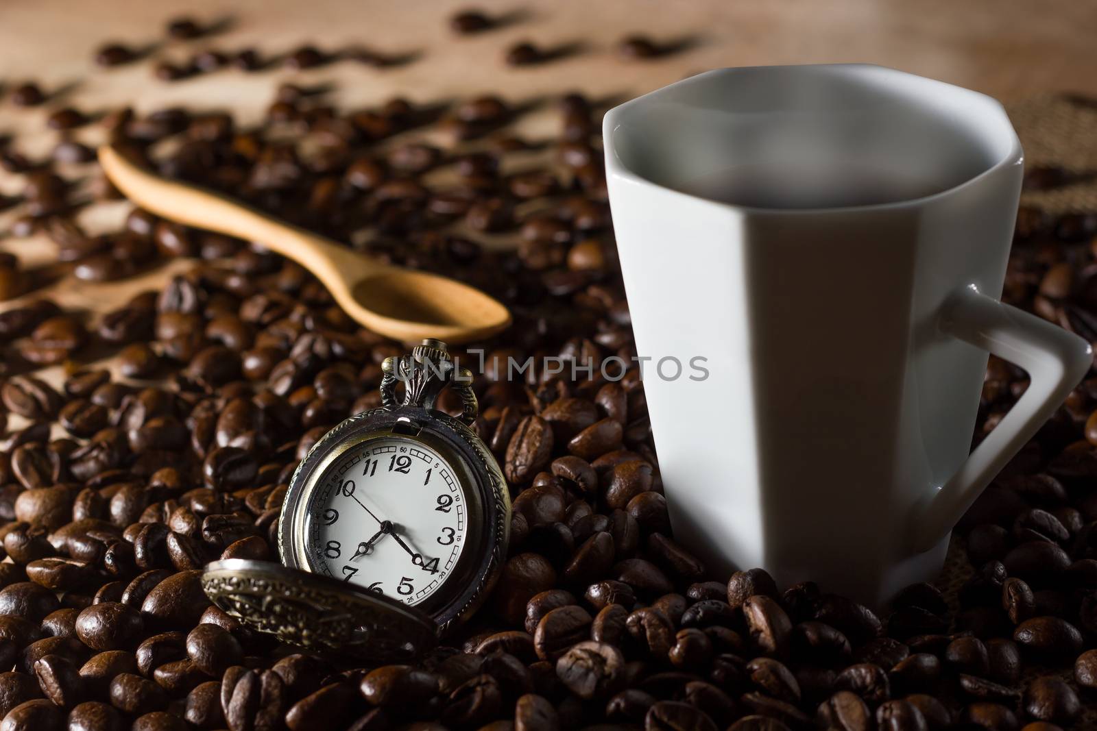 Low key coffee in the darkness. Roasted coffee beans on a wooden table with a glass of white ceramic and a wooden spoon placed behind an old watch.